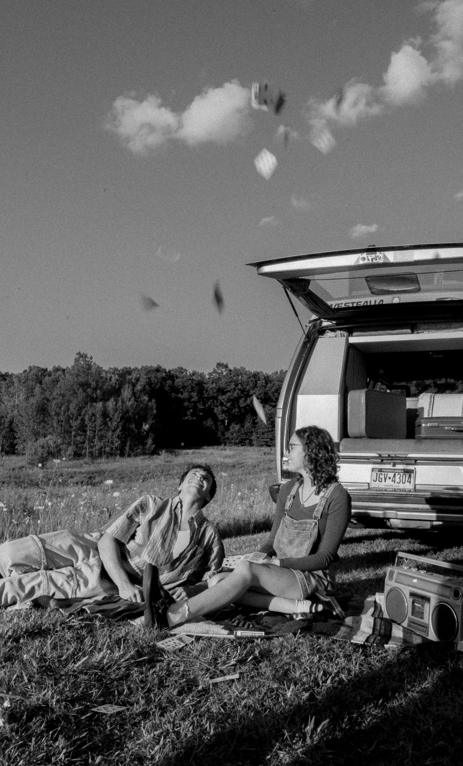 Two women relaxing on grass beside an open station wagon in a black and white outdoor summer scene.