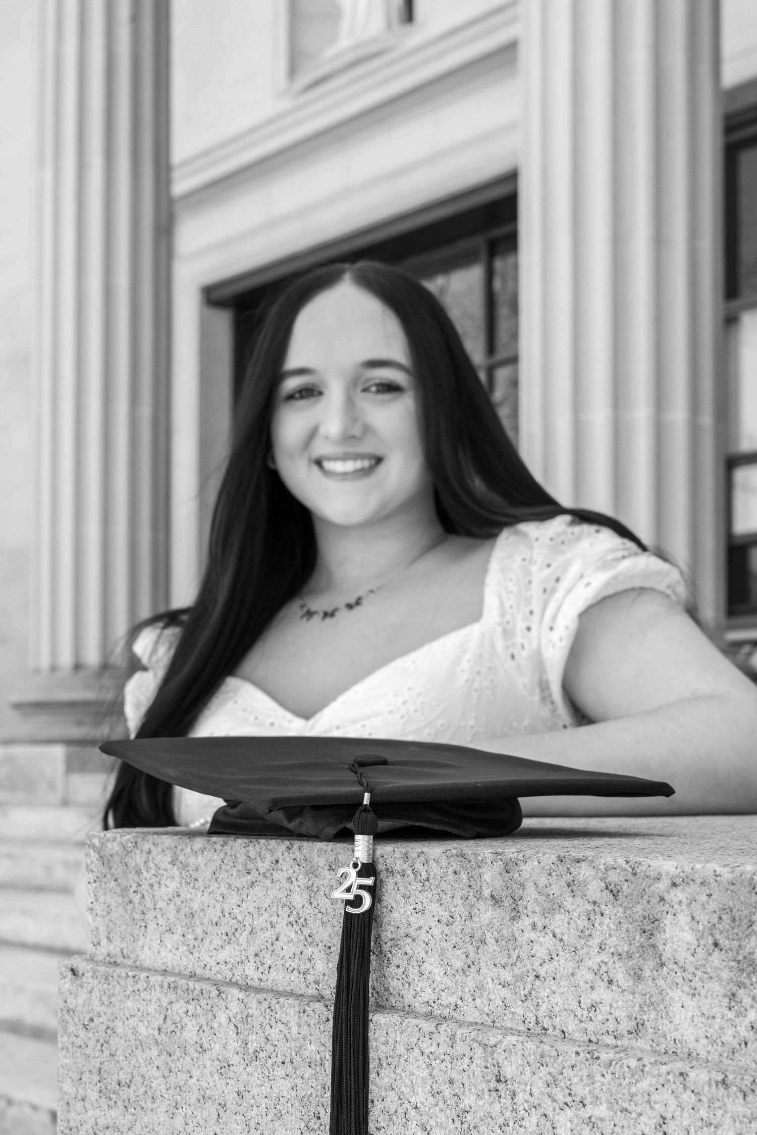 Black and white graduation photo of smiling young woman with long dark hair holding cap with 25 tassel on stone steps.