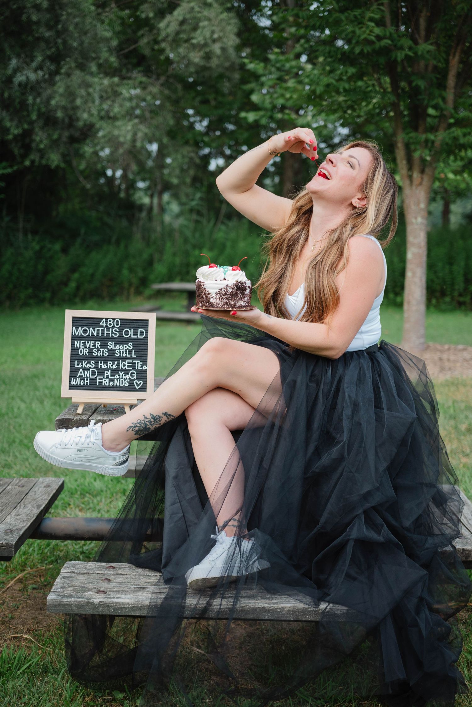 Woman in black tulle skirt eating birthday cake on picnic table with 480 months old chalkboard sign.