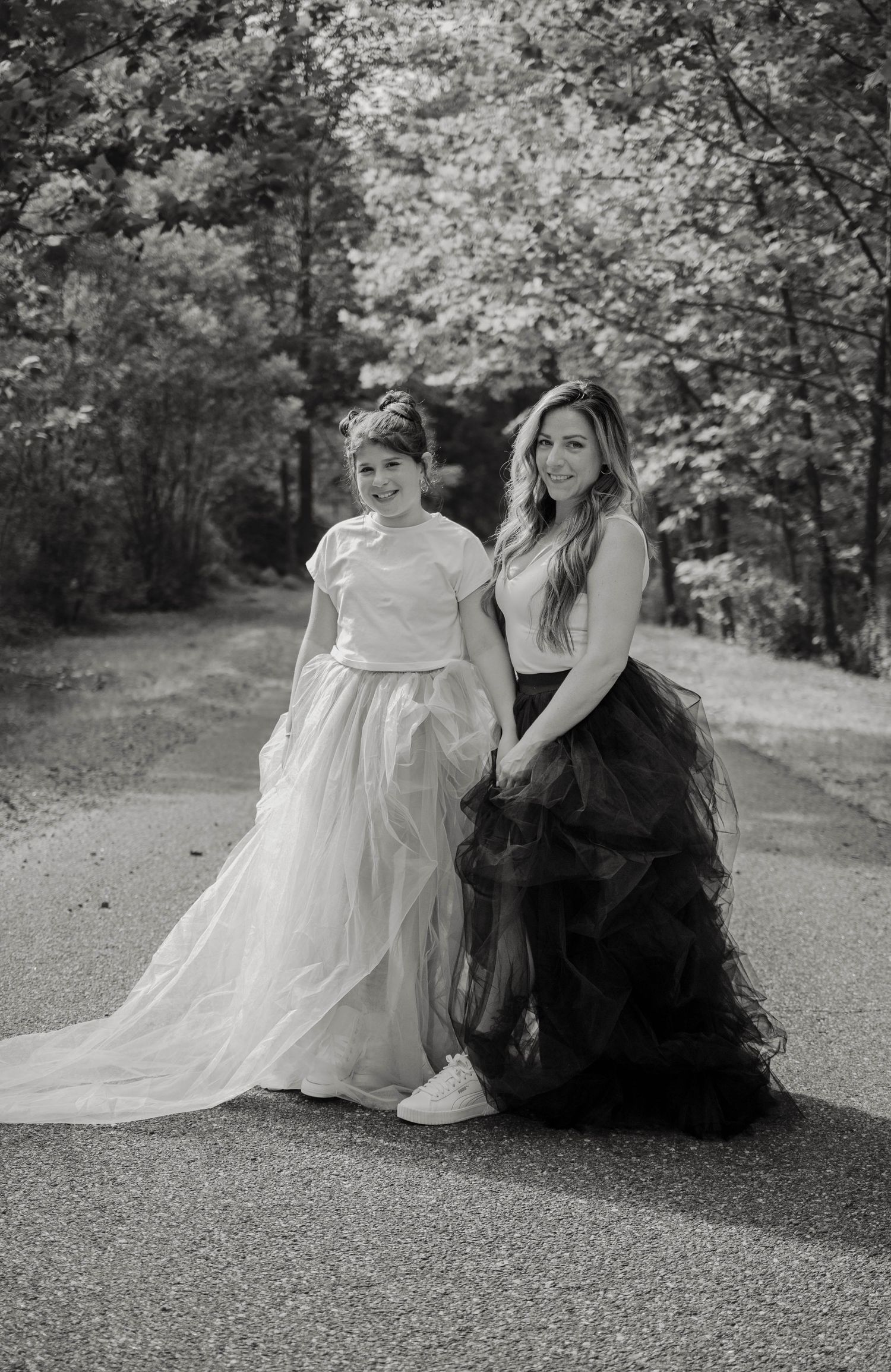 Two women pose on a path in black and white, wearing tulle skirts and white tops, surrounded by trees.