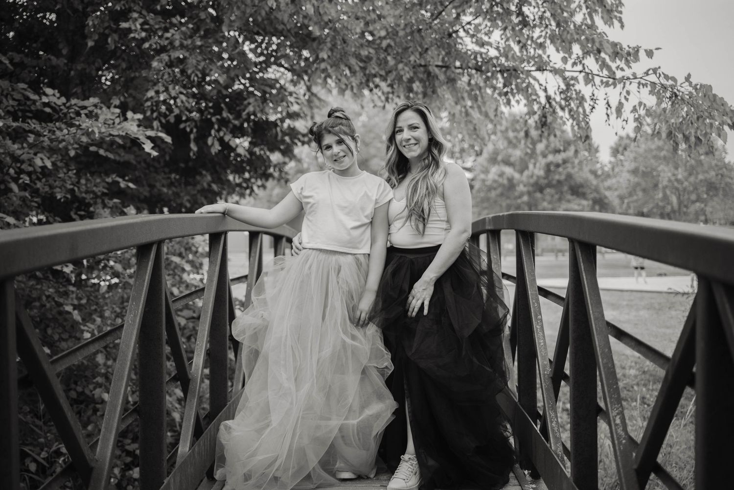 Two smiling women in tulle skirts pose together on a wooden bridge surrounded by lush green trees.