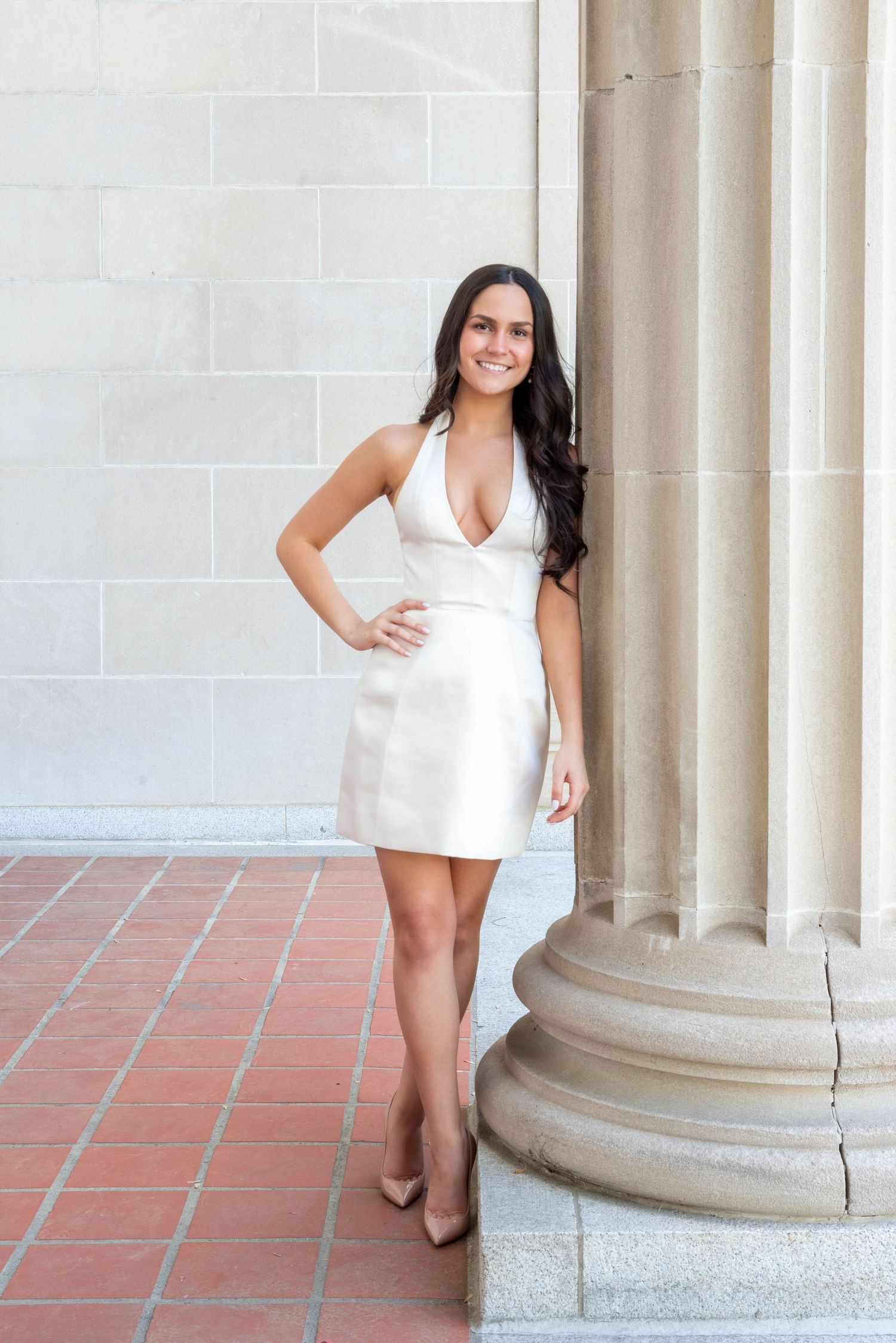 Young woman in white halter mini dress and nude heels posing by a classical stone column on a brick floor.