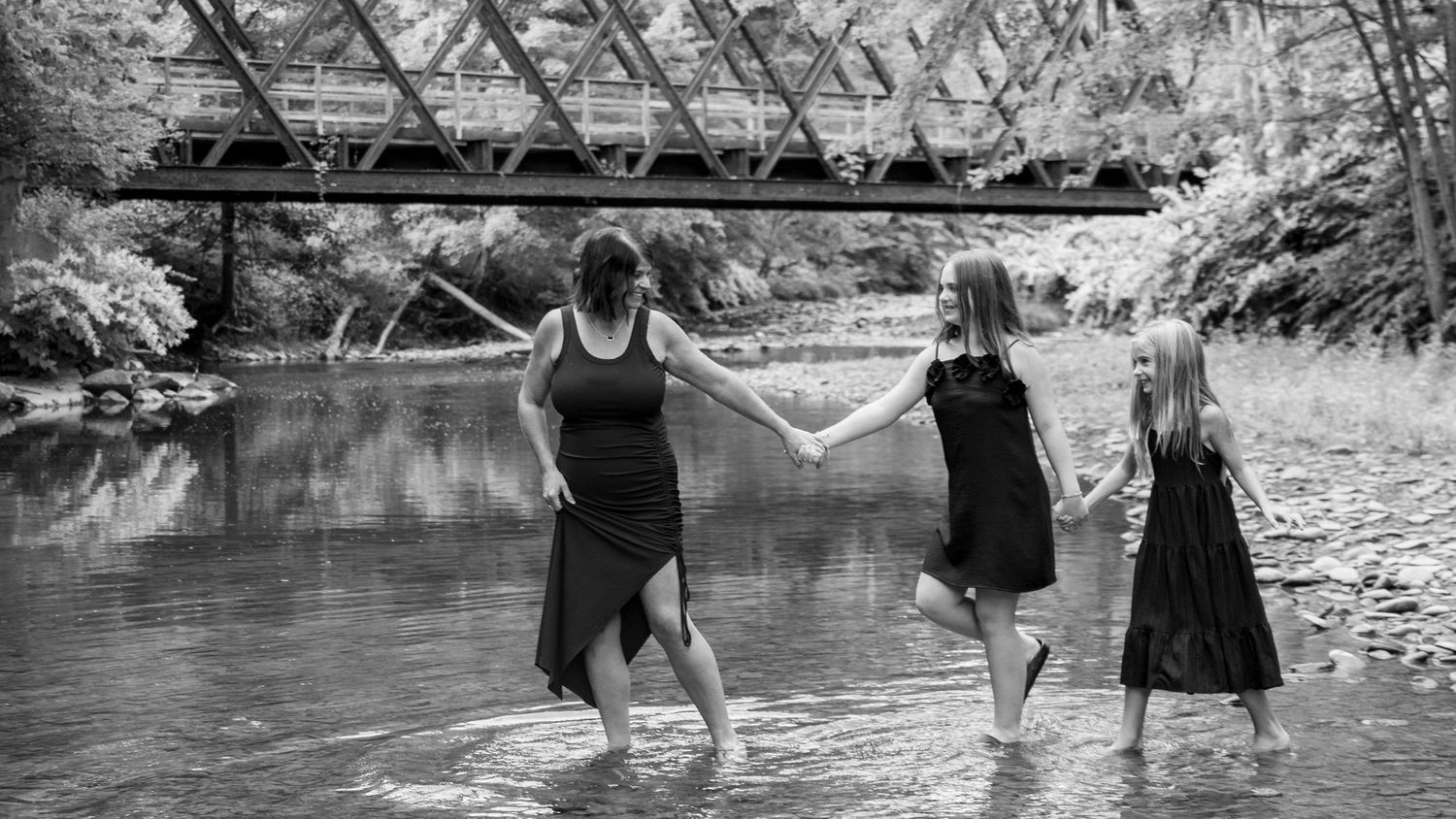 Three girls holding hands wade in a shallow river near a bridge in a black and white outdoor photo.