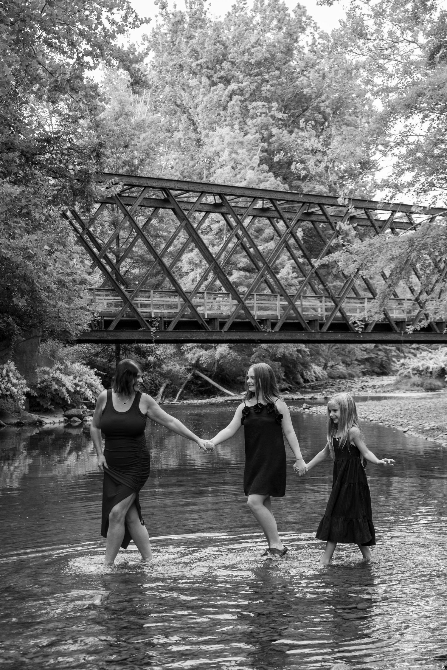 Three women in black dresses hold hands wading in a creek near a metal bridge, black and white photo.