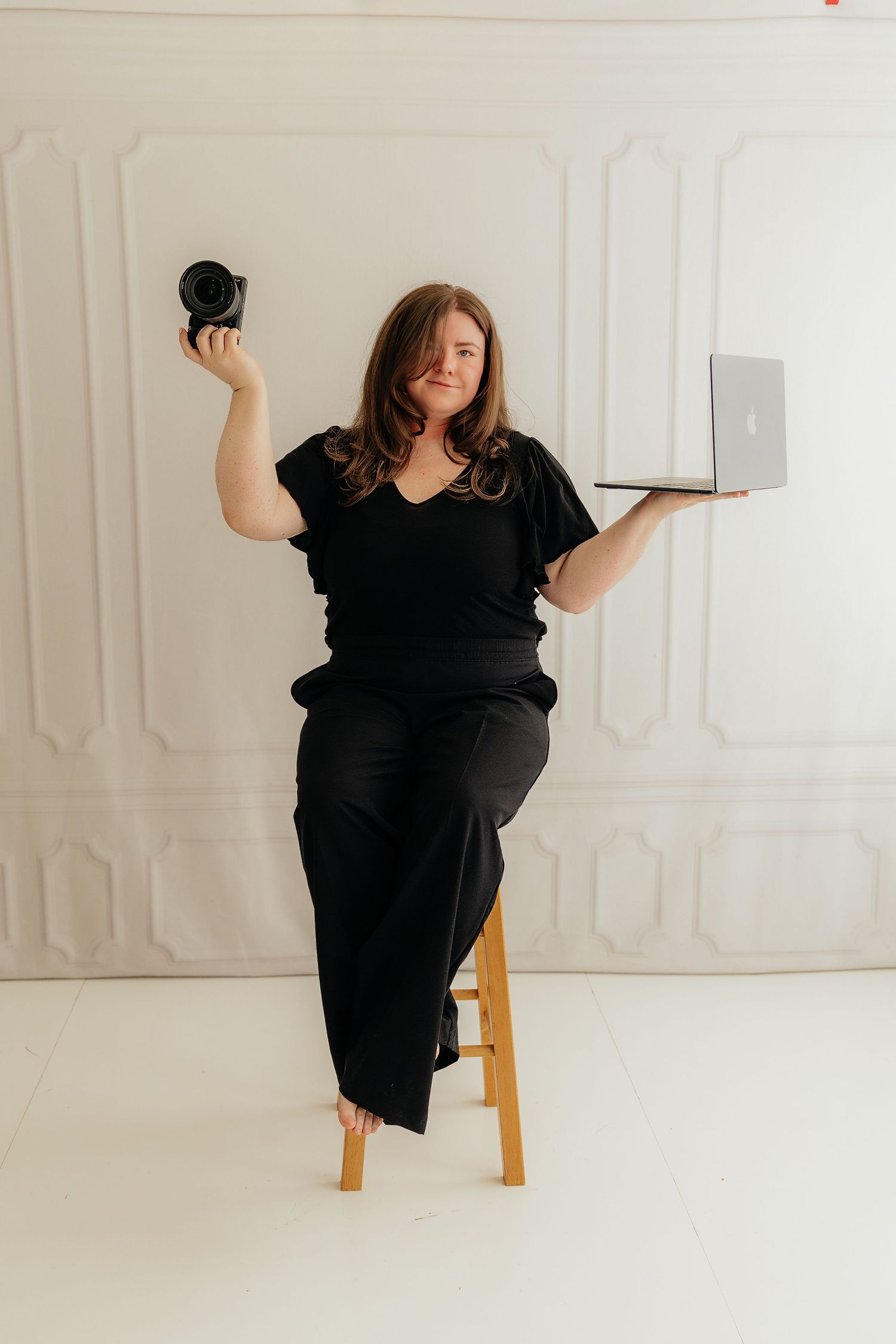 Woman in black outfit holding a camera and laptop while seated on a wooden stool against a white wall.