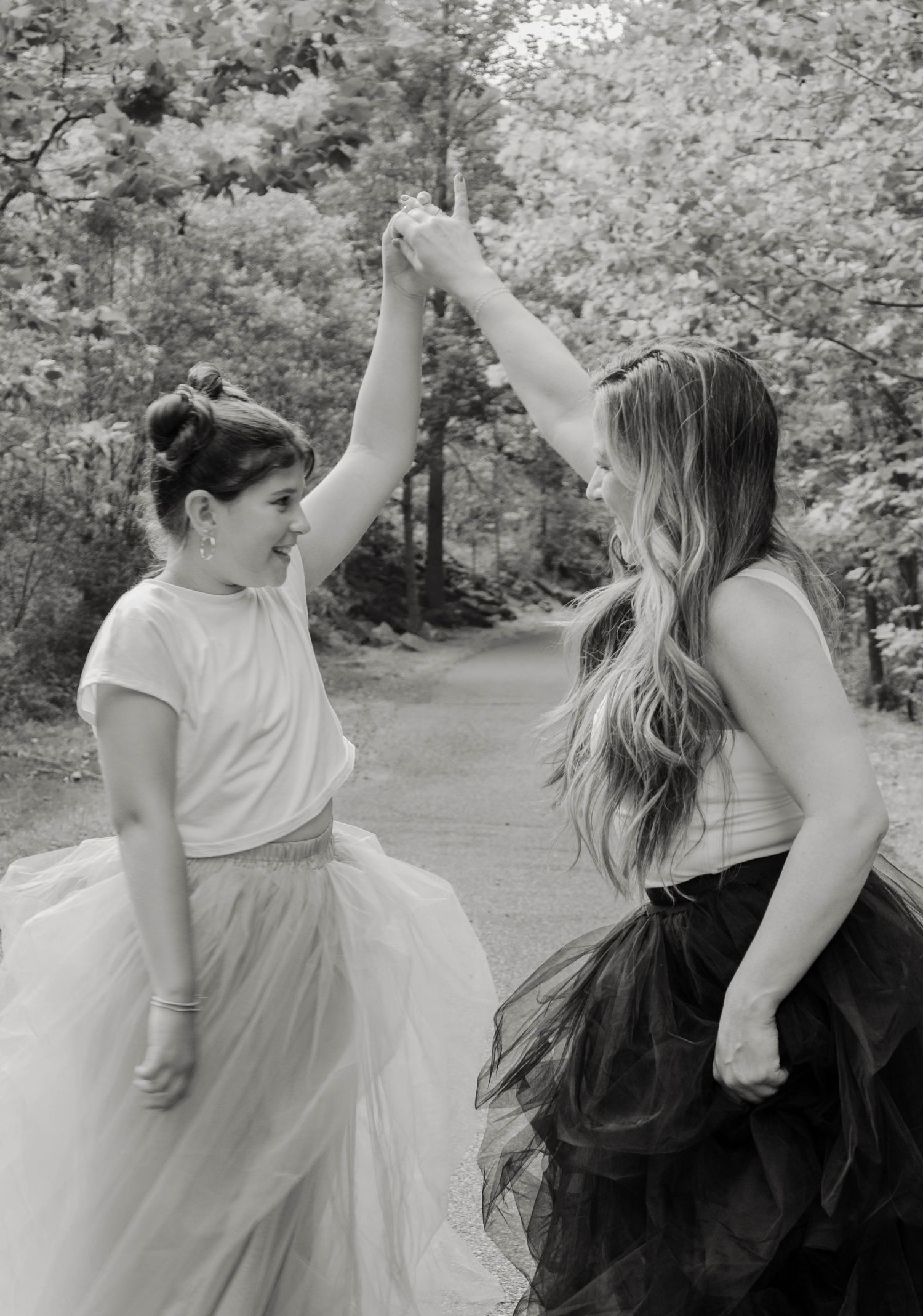 Two women in tulle skirts hold hands and twirl on a tree-lined path in a black and white photo.