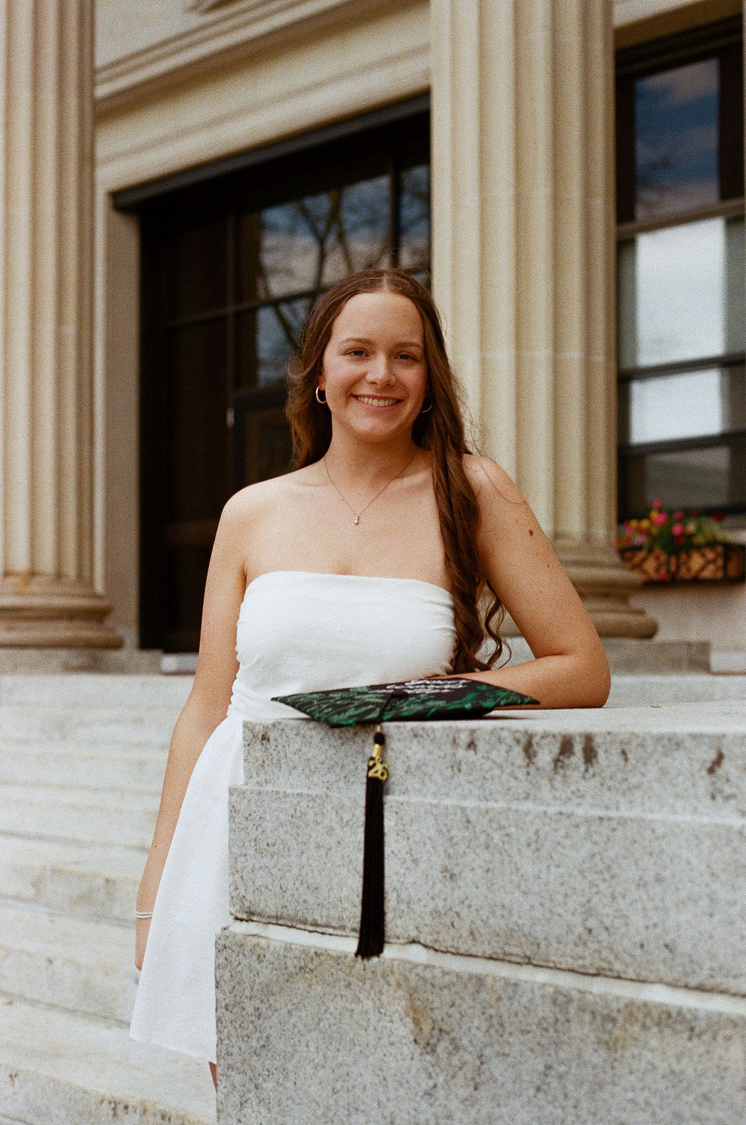 Young woman in white strapless dress holding graduation cap smiles on stone steps of a classical building.