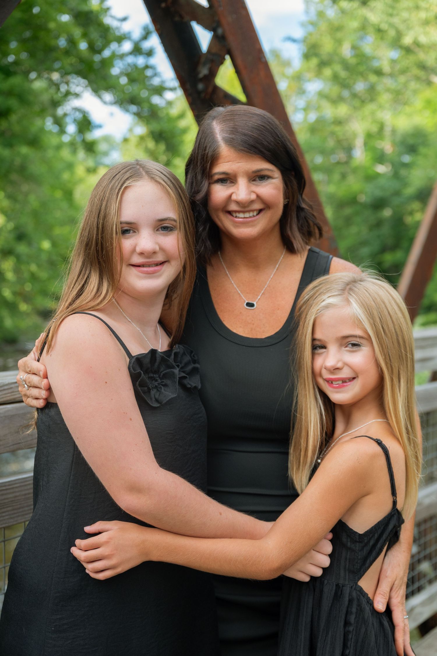 A smiling woman poses with two blonde girls in matching black dresses on a wooden bridge surrounded by green trees.