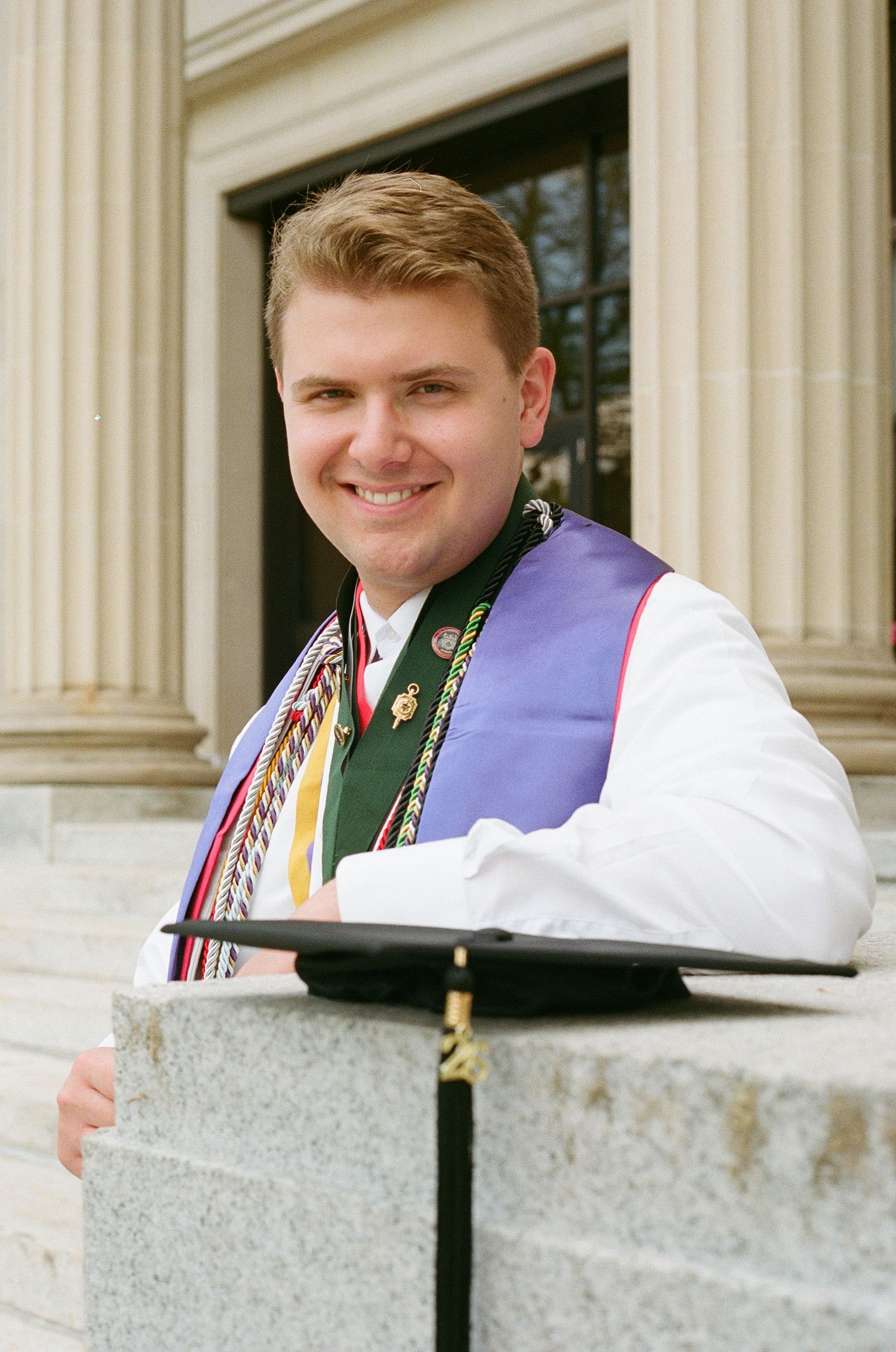Smiling young man in graduation regalia with colorful stoles and cap on college campus steps.