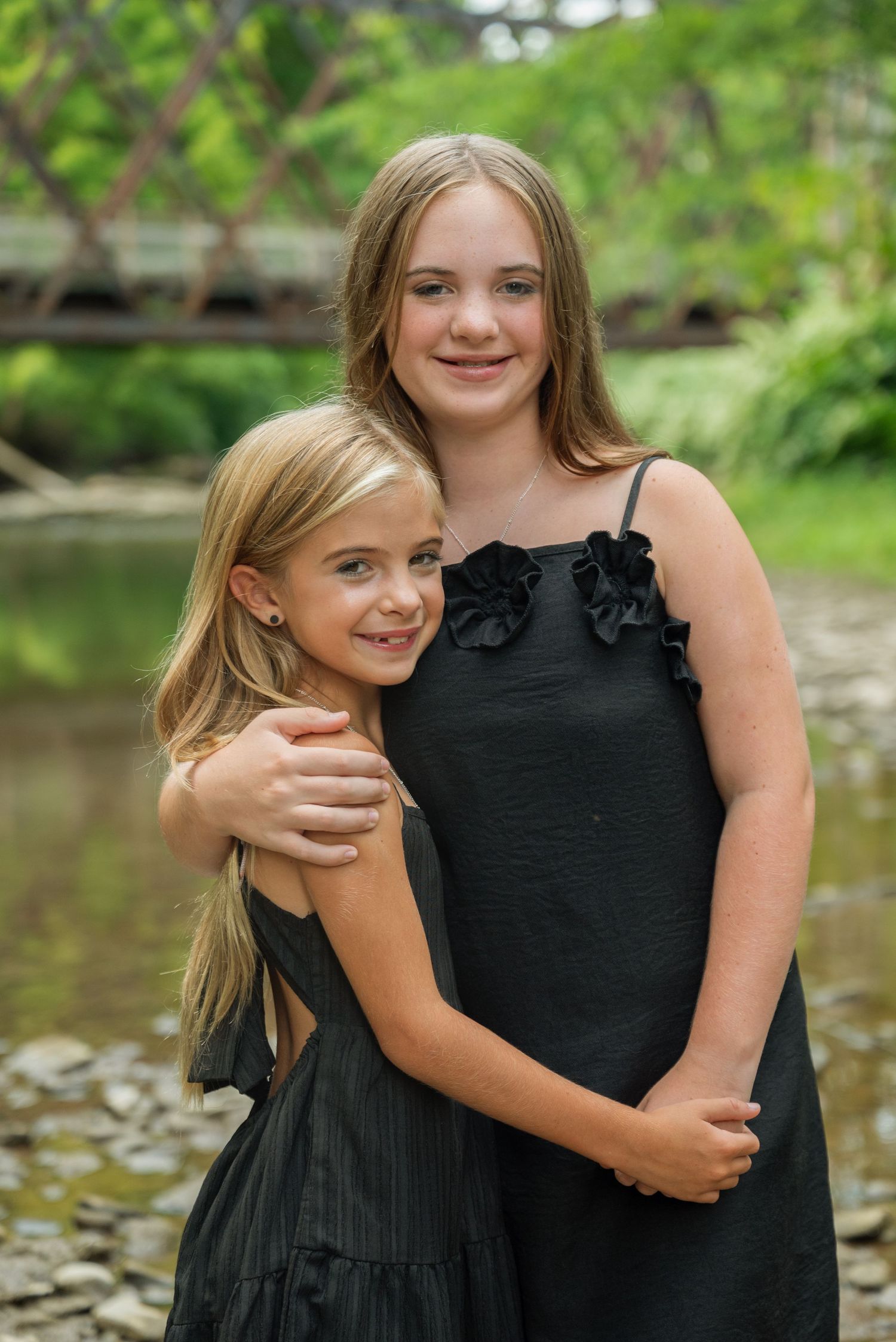 Two young girls in matching black dresses embrace outdoors near a creek with a wooden bridge in the background.