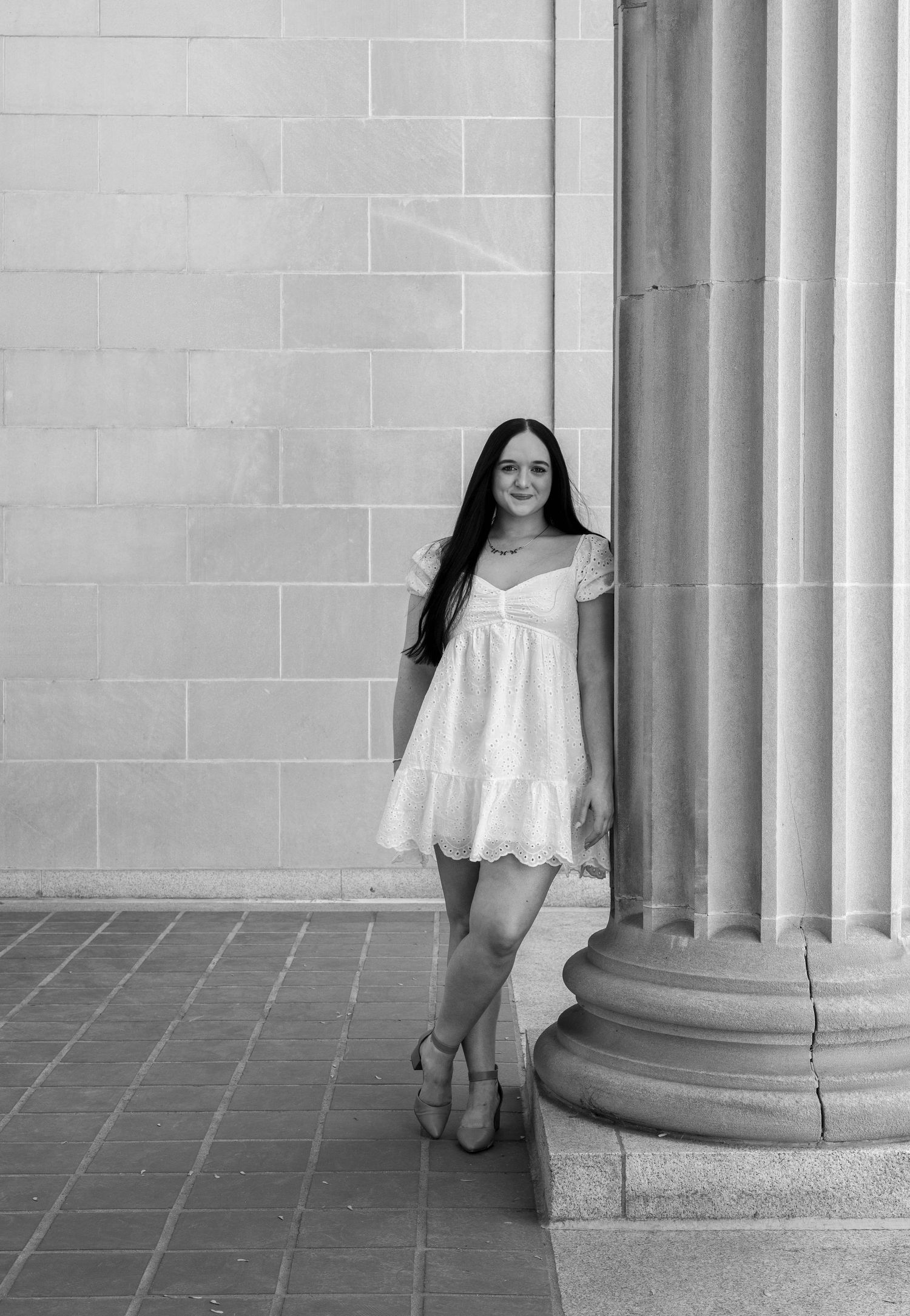 Young woman in white dress leaning against a stone column, elegant black and white portrait.