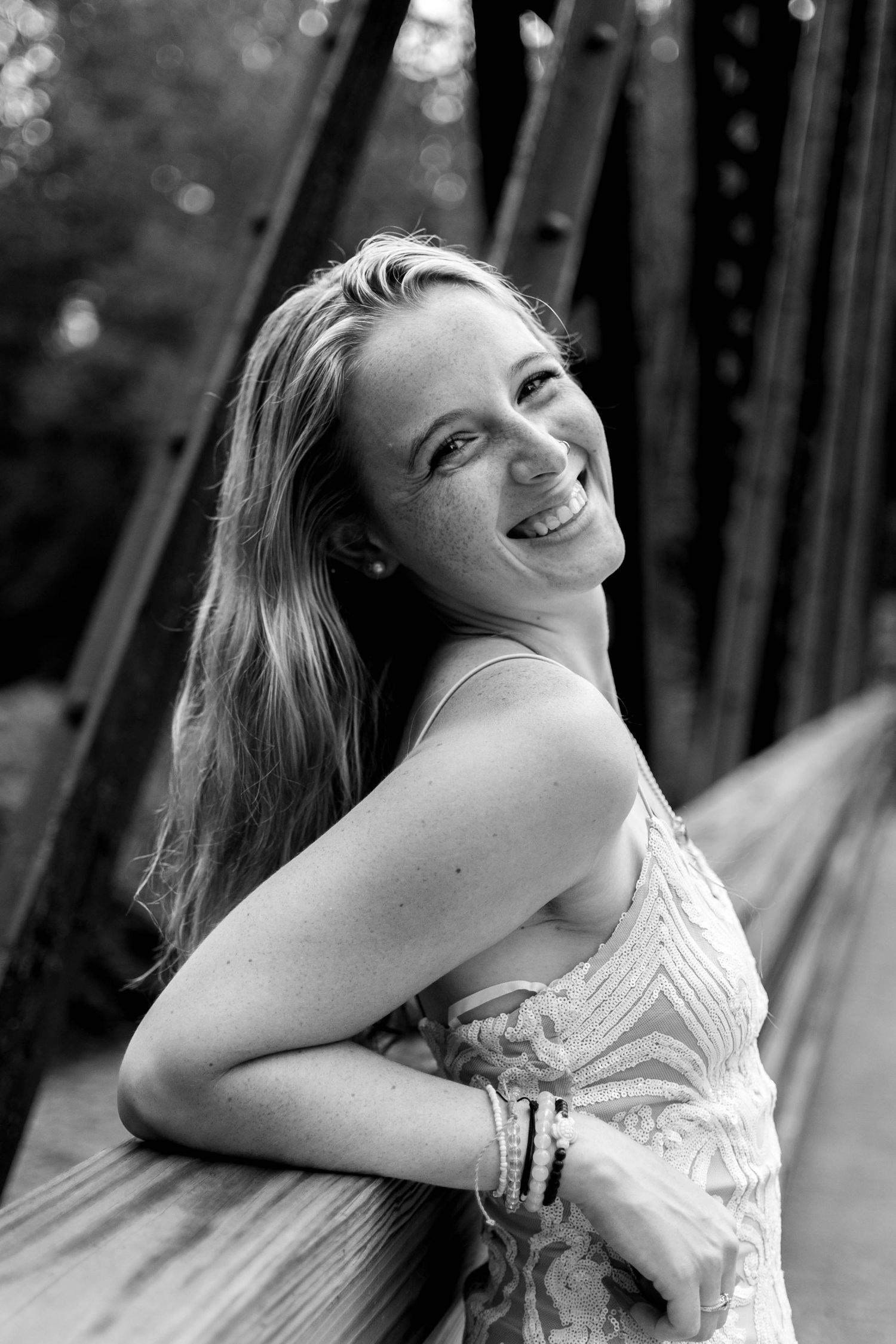 Black and white portrait of smiling teenage girl with freckles leaning on bridge railing outdoors.