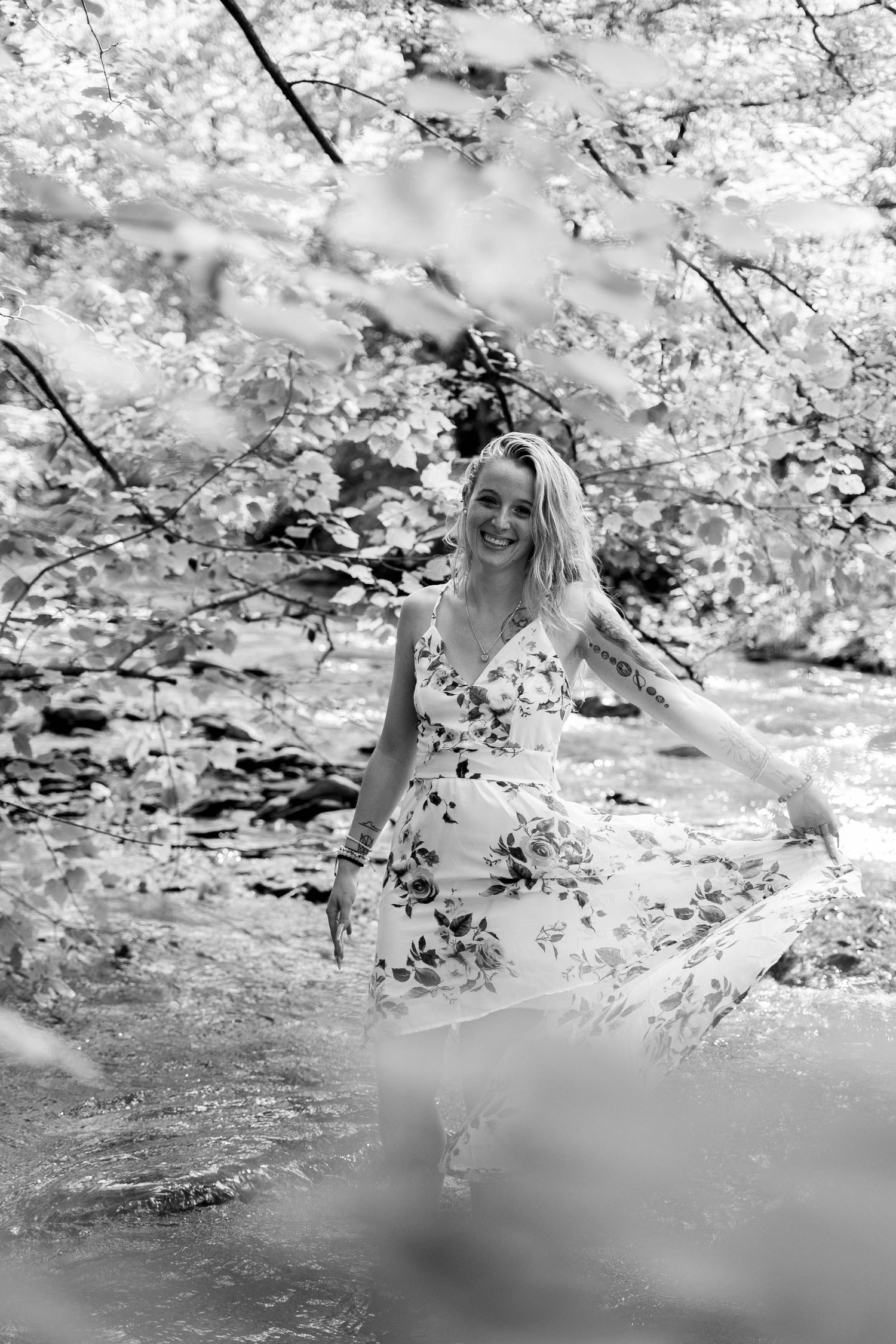 Smiling blonde woman in floral dress dancing by a sunlit forest river, black and white photo.