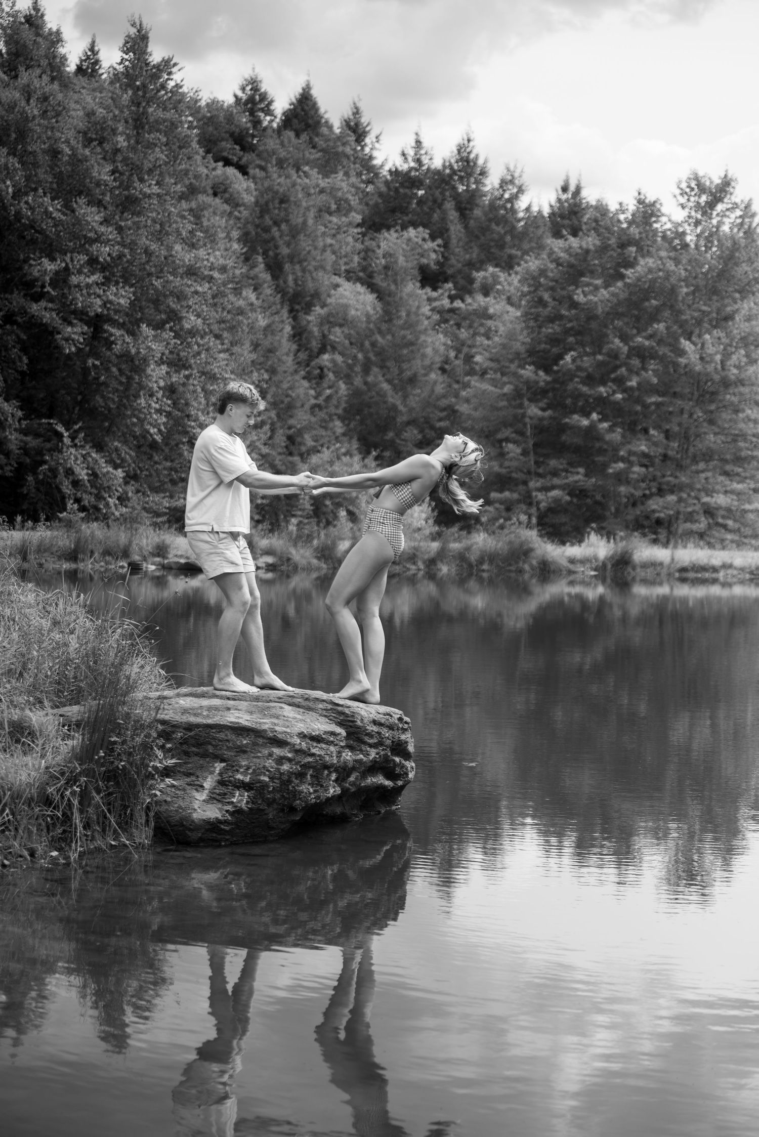 Couple holding hands on a rock by a serene lake surrounded by lush forest in a romantic black and white photo.