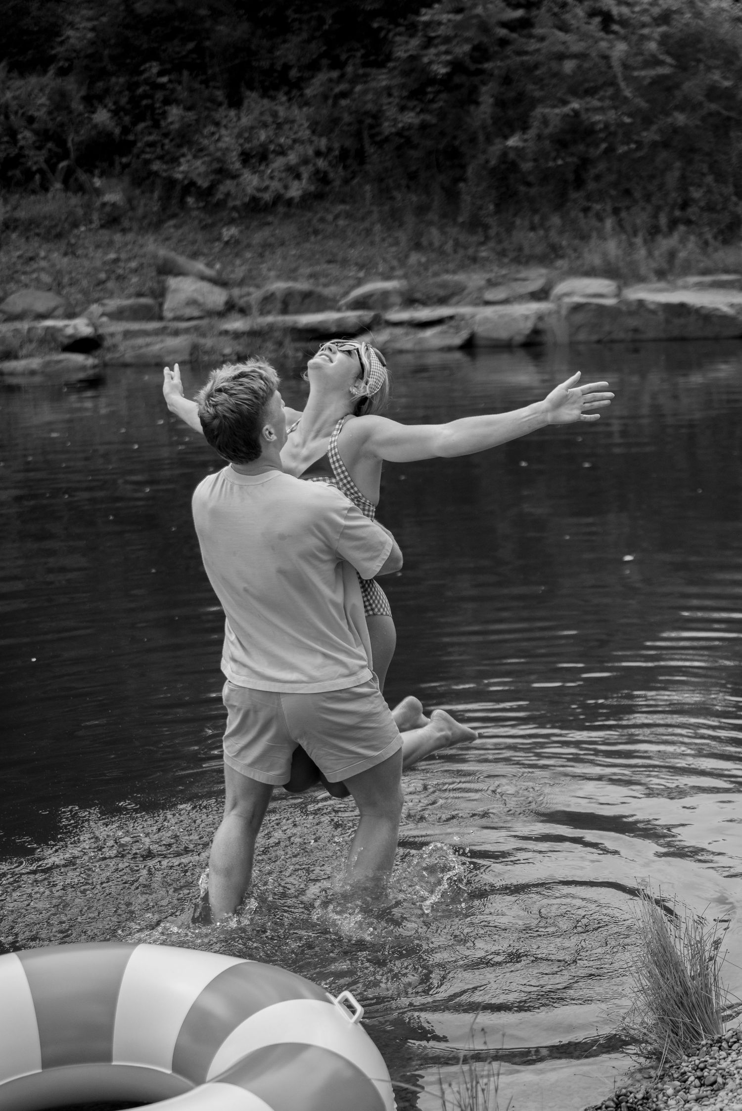 Black and white photo of a couple laughing and playing in a lake with a striped inflatable ring nearby.