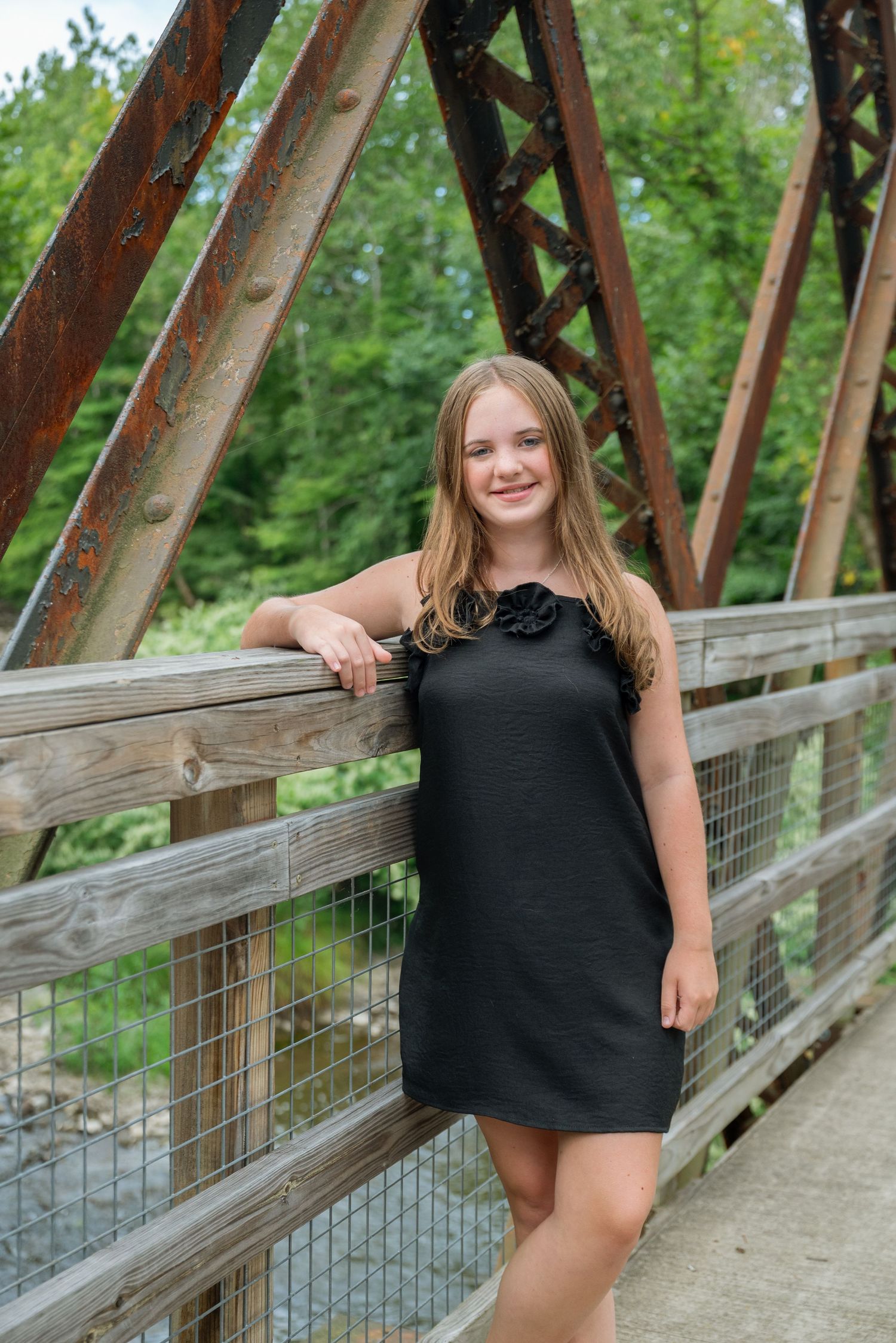 A smiling teenage girl in a black dress poses on a rustic wooden bridge with iron trusses surrounded by lush greenery.
