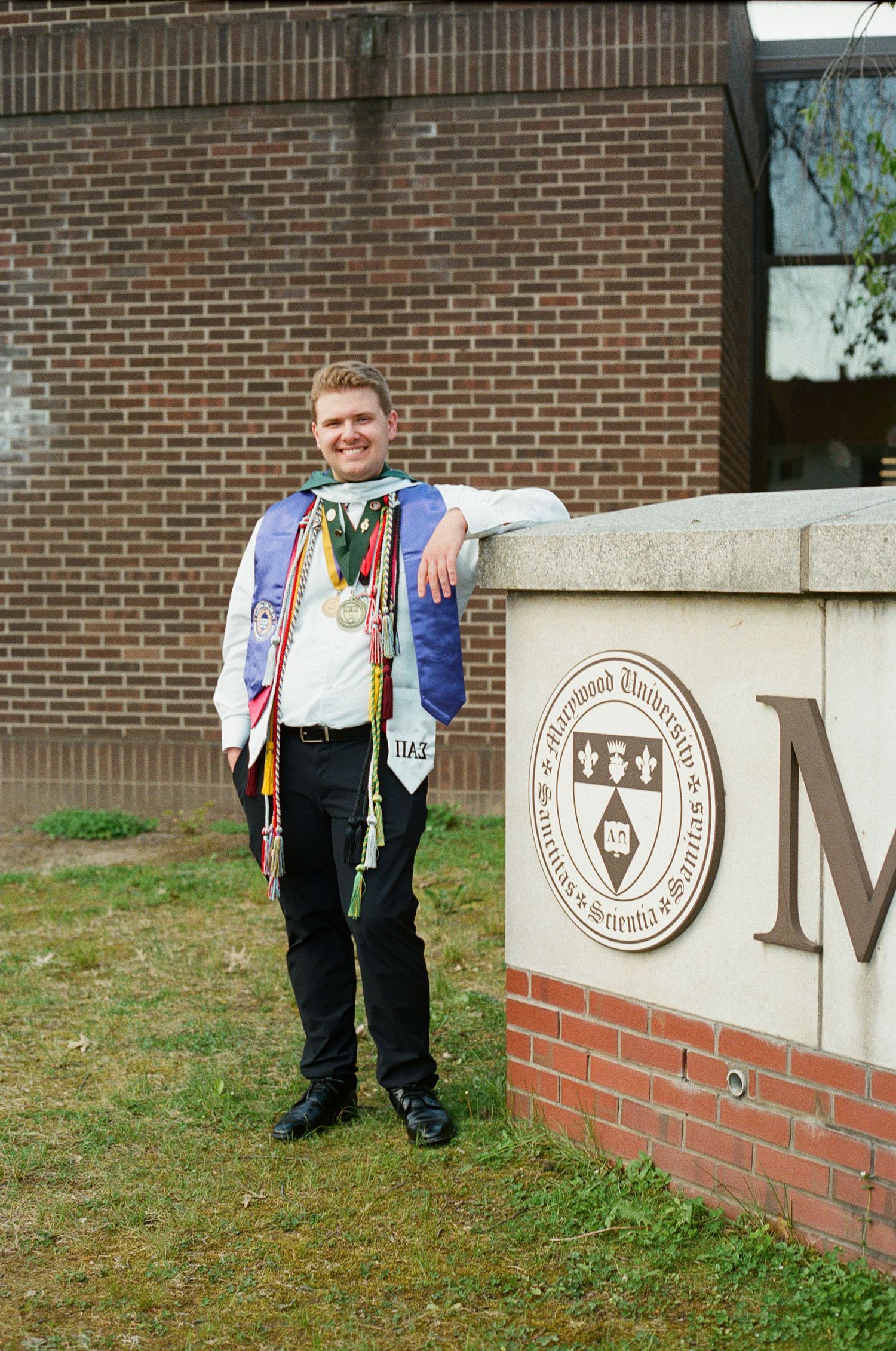 Smiling graduate posing beside a Marywood University sign wearing colorful honor cords and medals.
