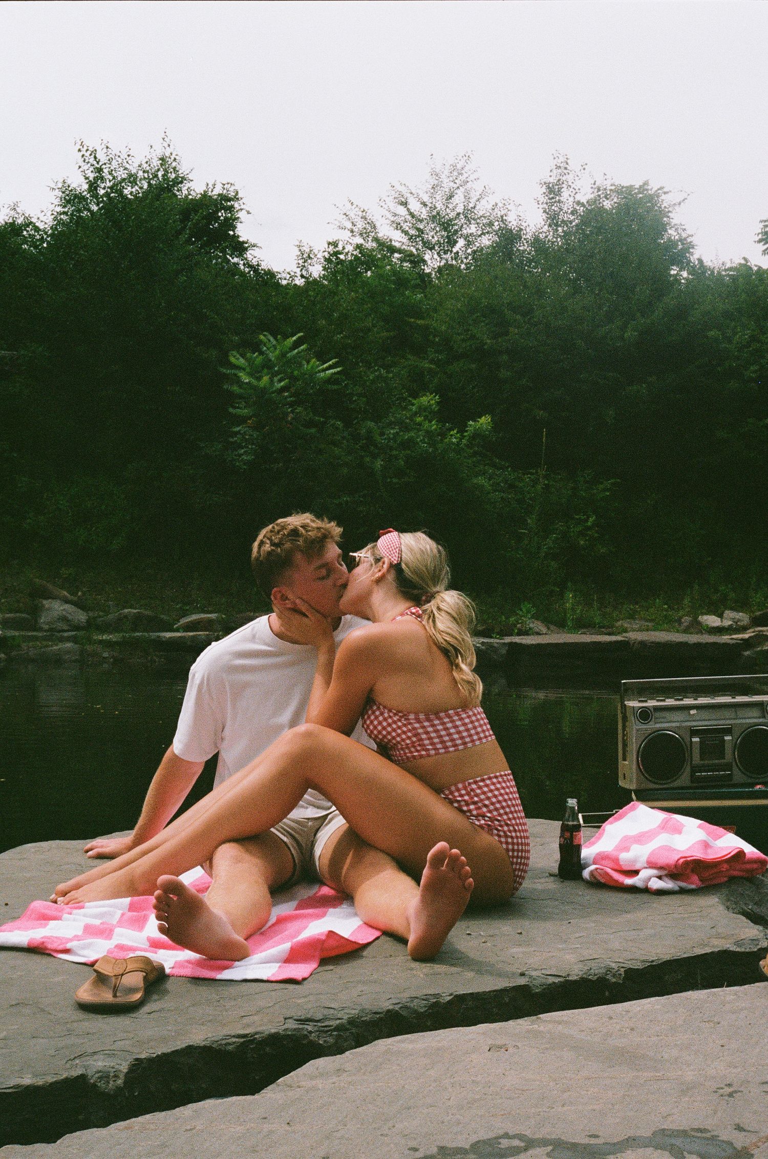 A couple shares a romantic kiss on a rocky lakeside, sitting on a pink checkered blanket with a boombox nearby.