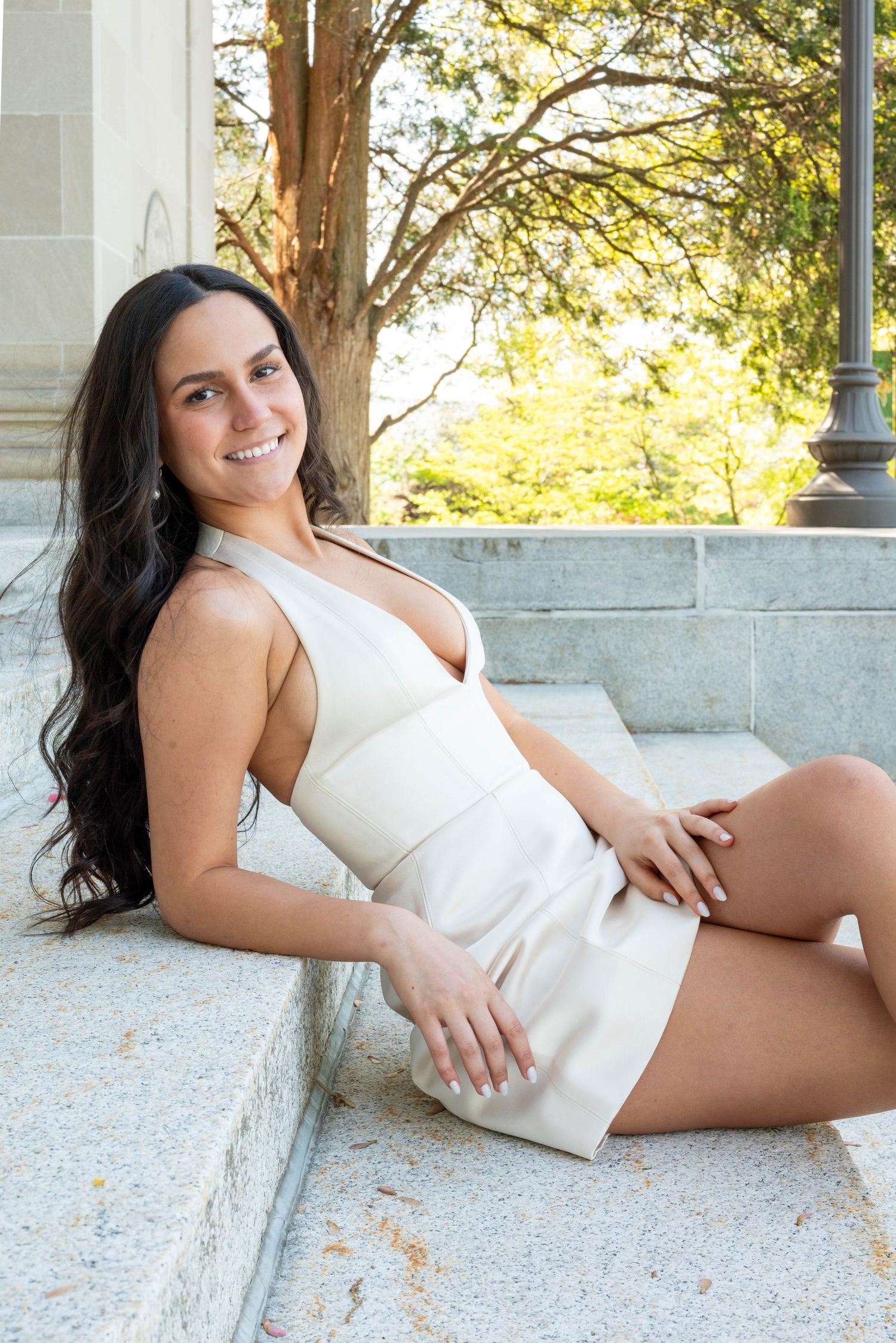 Smiling young woman with long dark hair sitting on stone steps wearing a white mini dress outdoors.