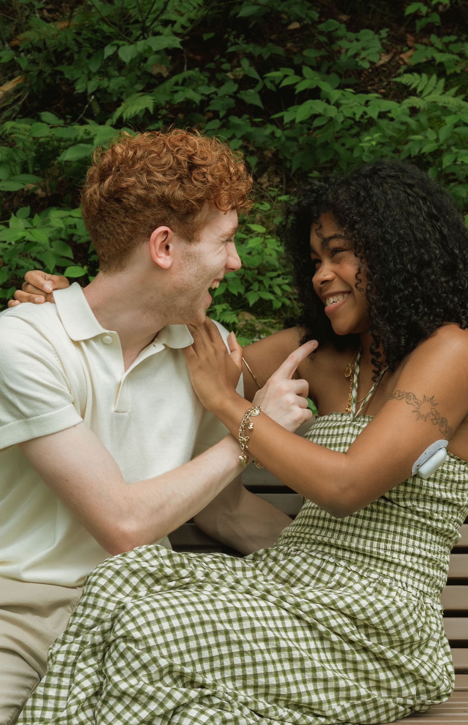 A joyful interracial couple laughs together on a bench surrounded by lush green foliage outdoors.