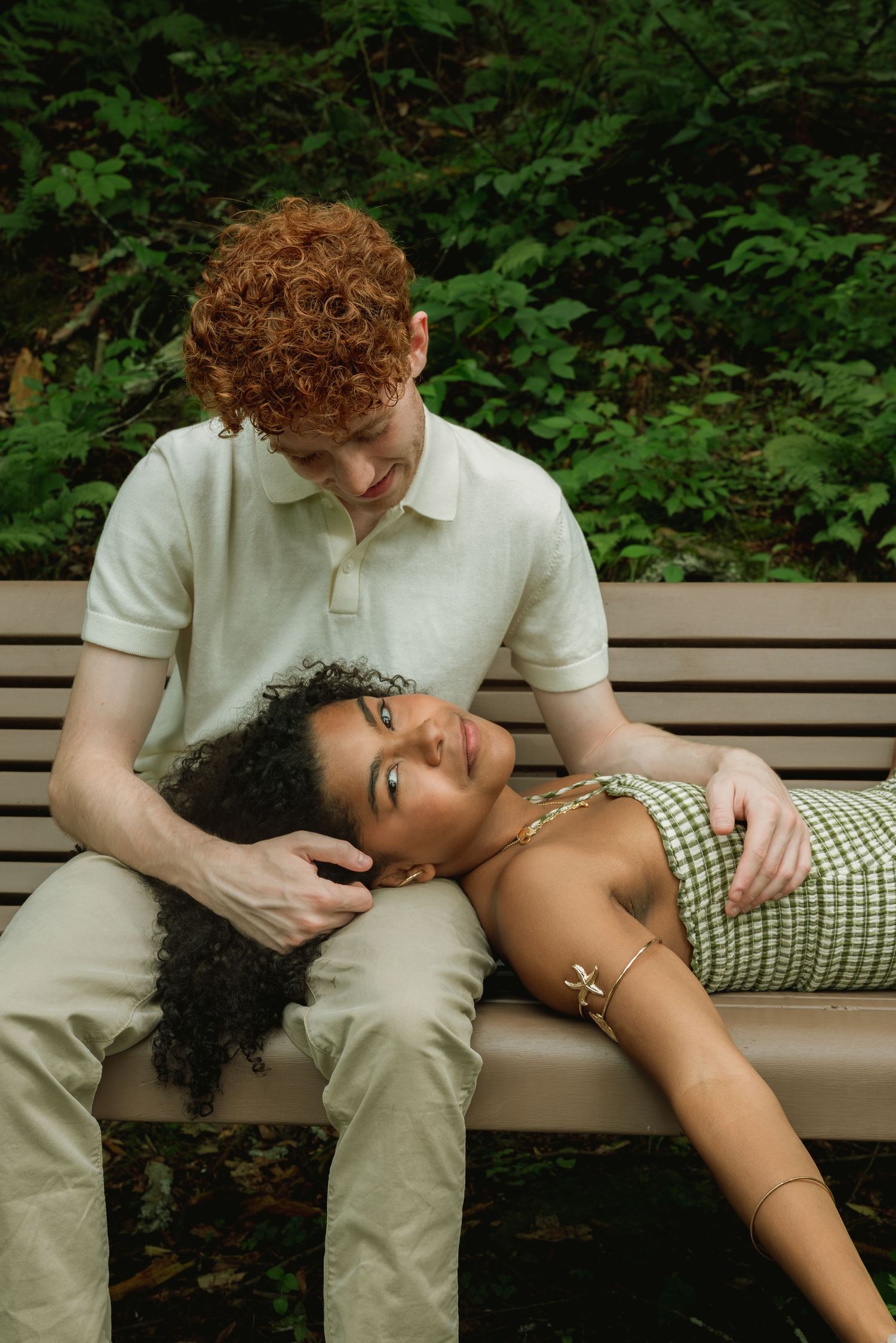 A couple relaxes on a park bench surrounded by greenery, woman lying in mans lap looking up tenderly.