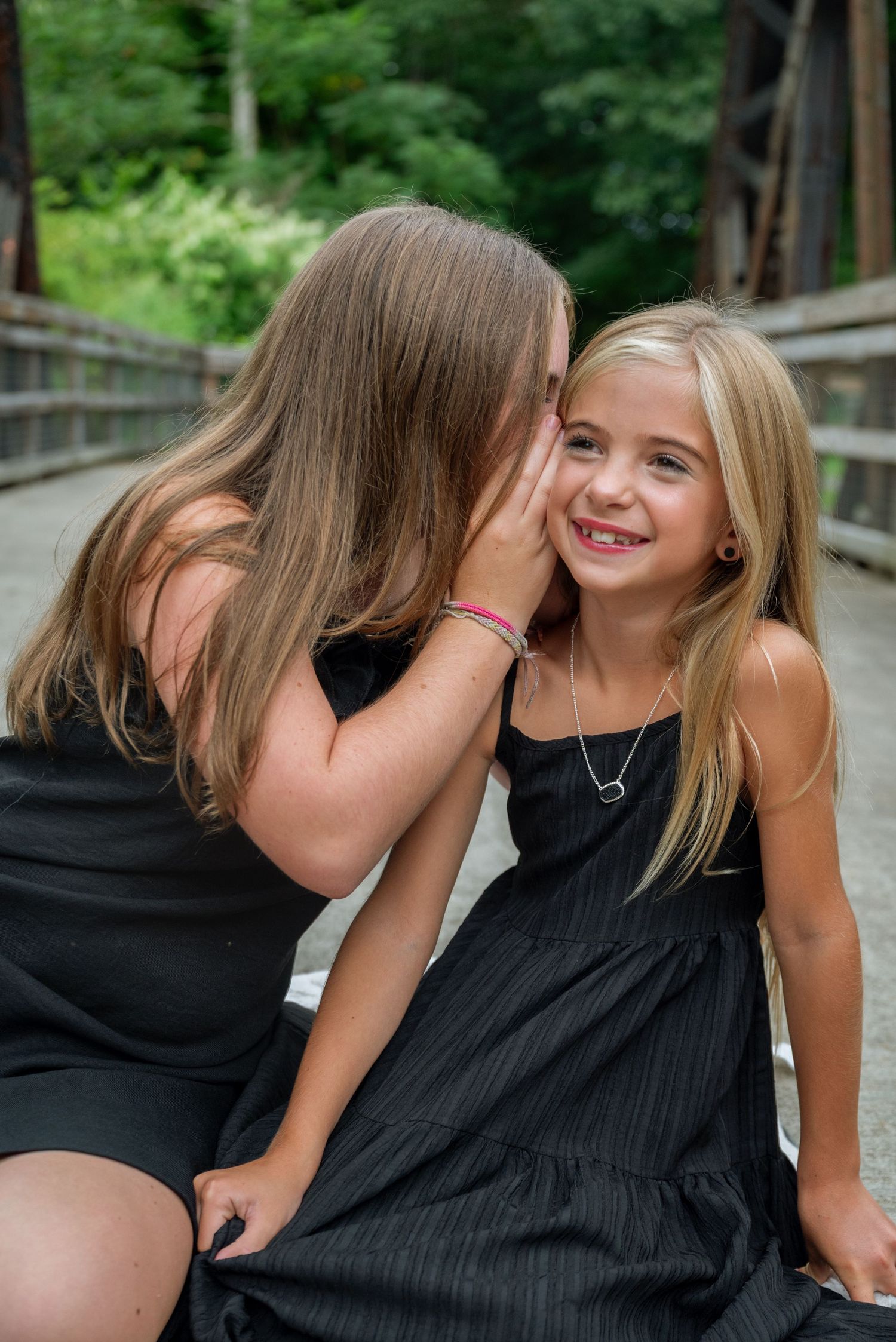 Two young girls in black dresses share a secret whisper on a bridge, one smiling joyfully.