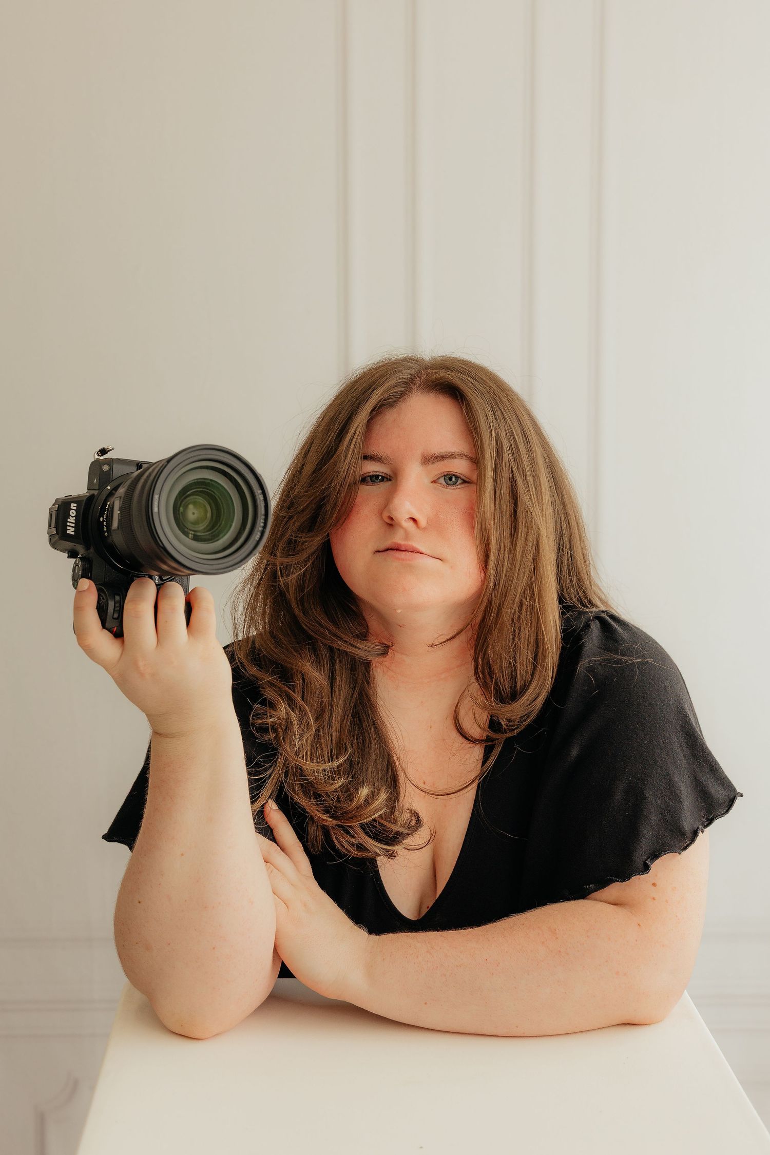 A woman with long brown hair holds a DSLR camera while leaning on a white table, wearing a black top.