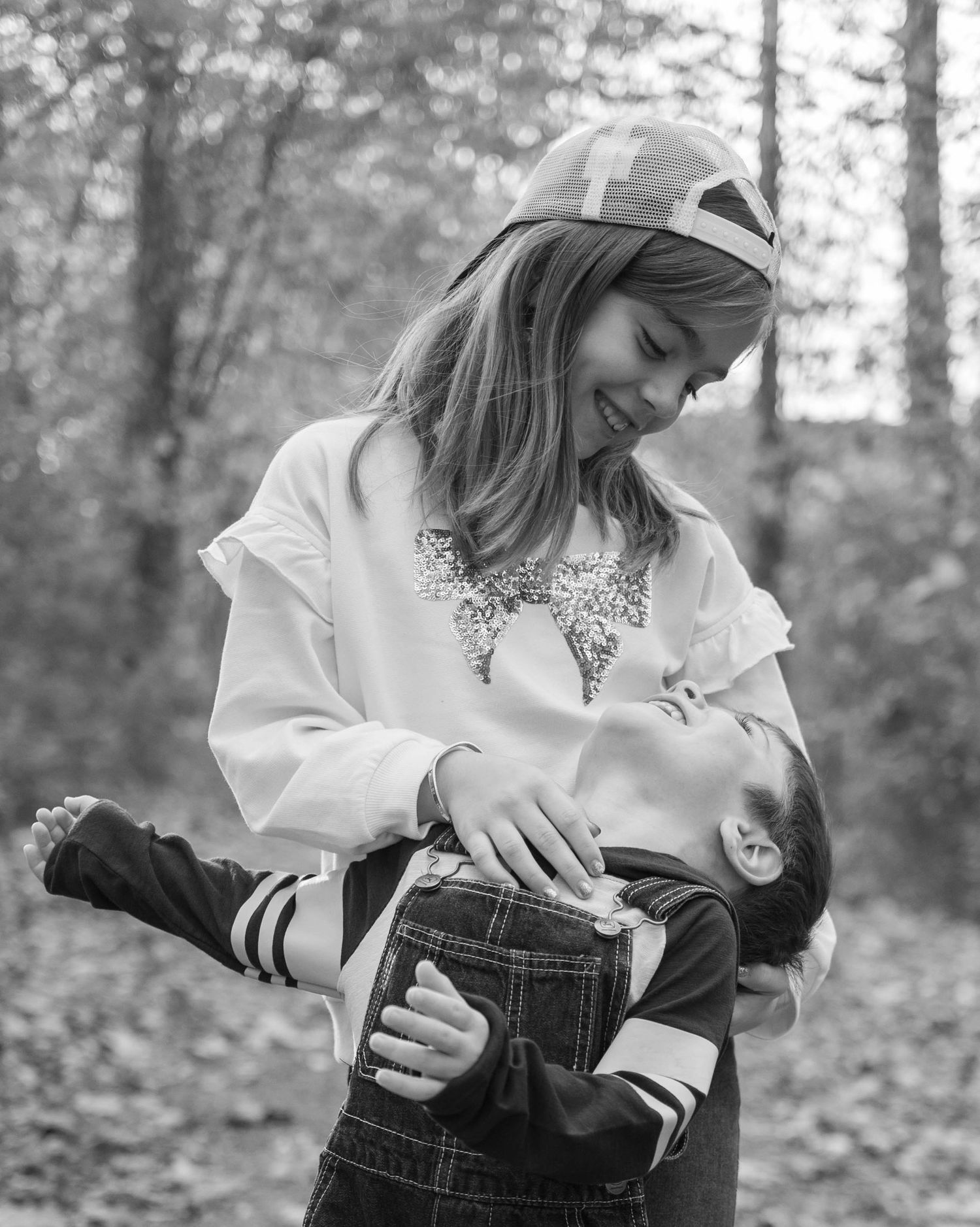 A smiling girl playfully dips her younger brother in a black and white outdoor forest portrait.