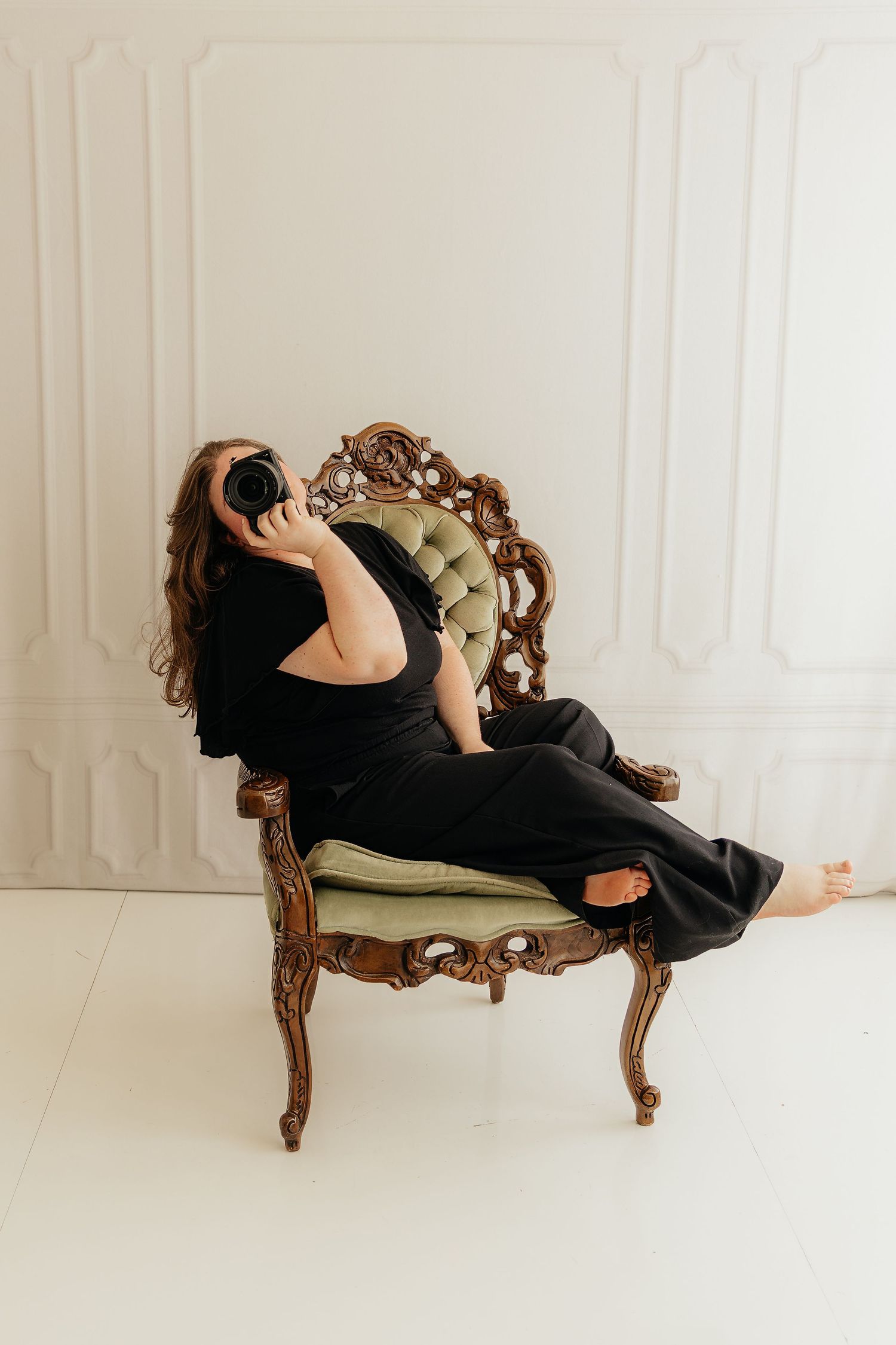 Woman in black outfit reclines on ornate vintage chair holding camera in bright white studio.