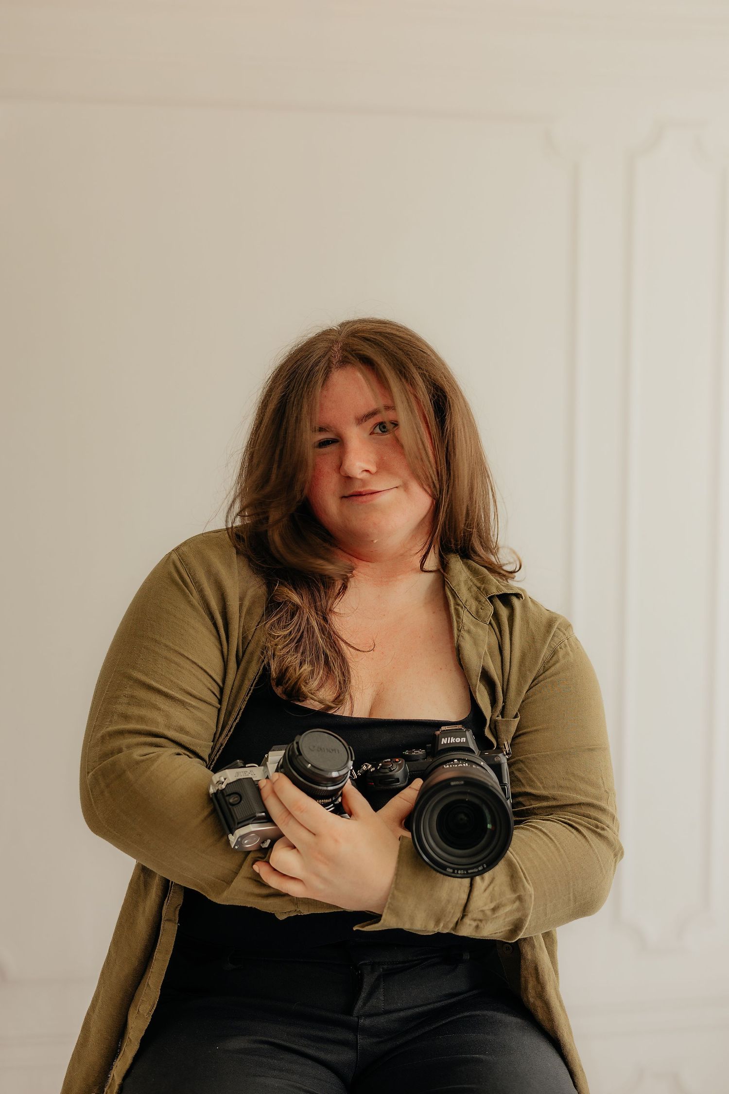 Woman photographer holding a Nikon camera, wearing olive jacket, posing against white wall backdrop.