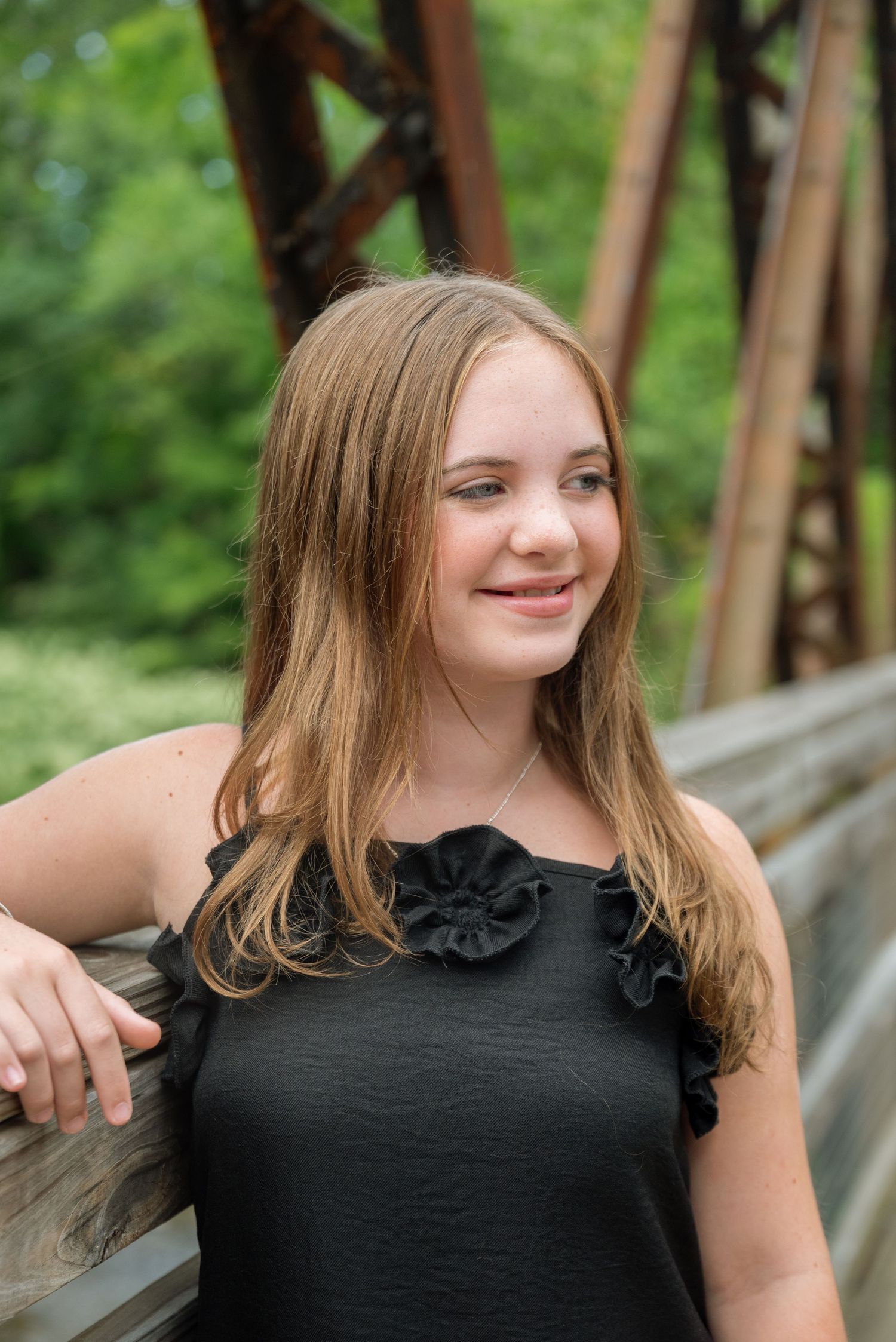 Teen girl with long blonde hair smiling in black ruffled top on a rustic bridge with green trees.