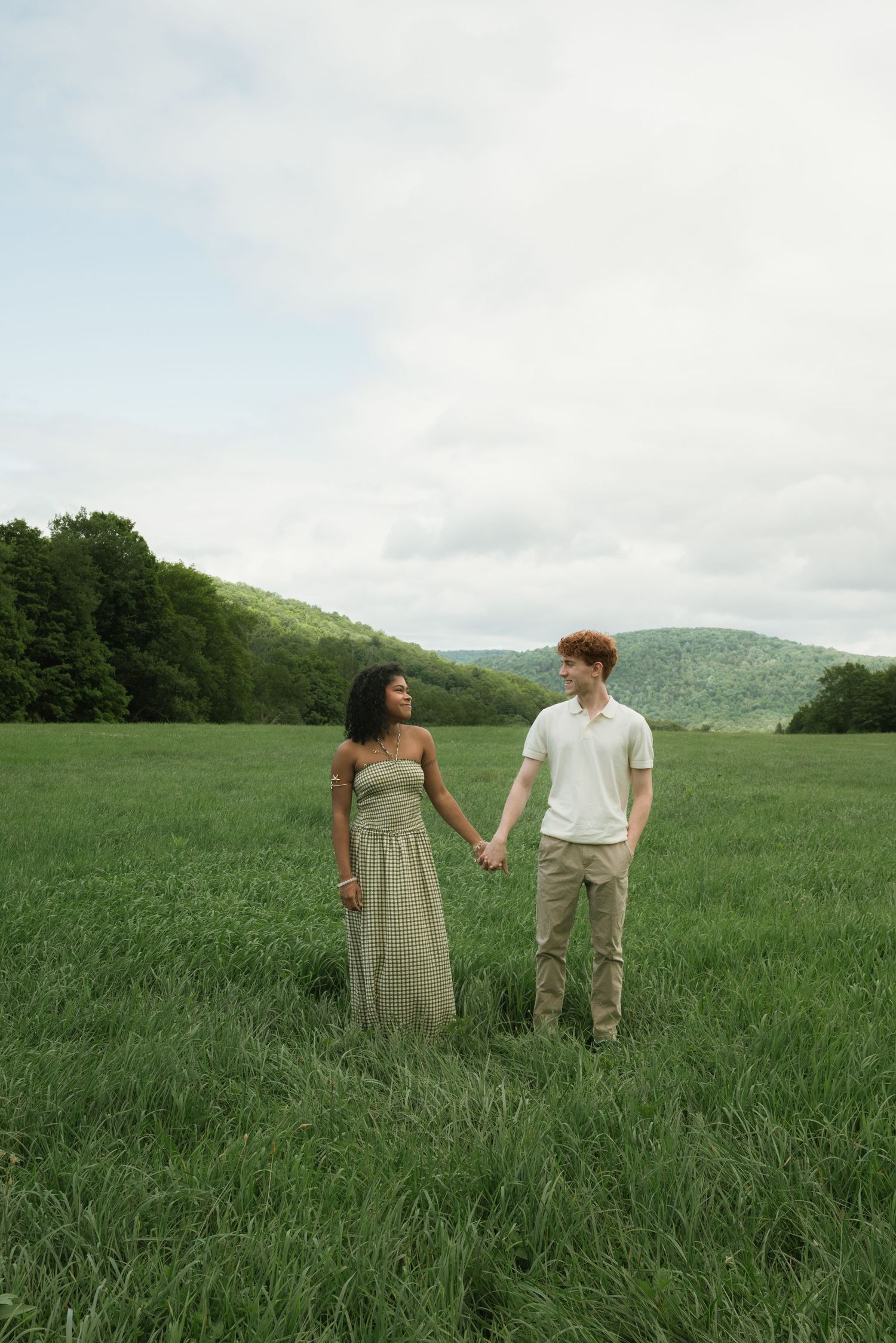 A couple holds hands in a lush green meadow with rolling hills and trees in the background under a cloudy sky.