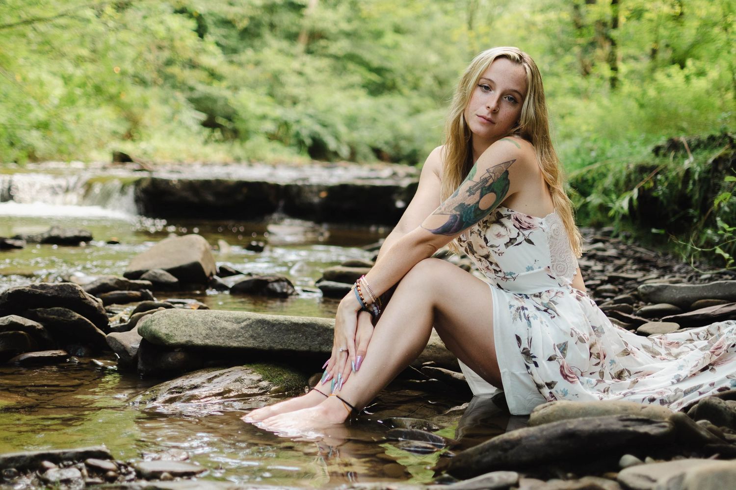 Tattooed blonde woman in floral dress sitting on rocks by a woodland stream.