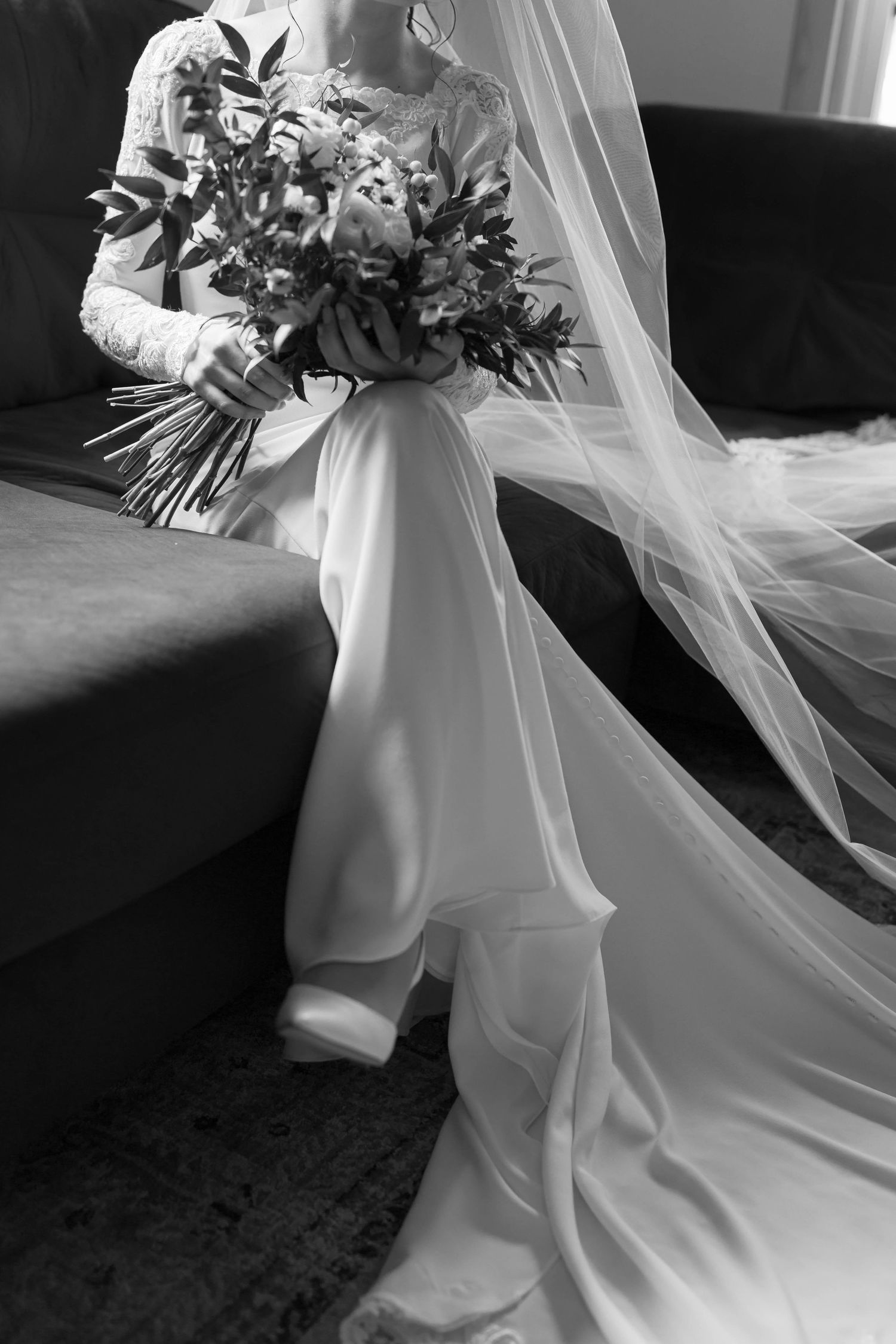 Black and white bridal portrait of bride sitting on sofa with long flowing veil and bouquet draped across her lap, elegant wedding detail.