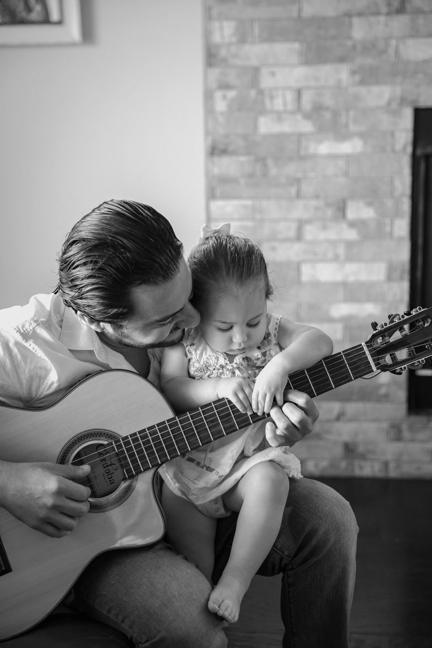 Black and white candid photo of a father playing acoustic guitar while a toddler sits on his lap touching the strings, cozy family moment in