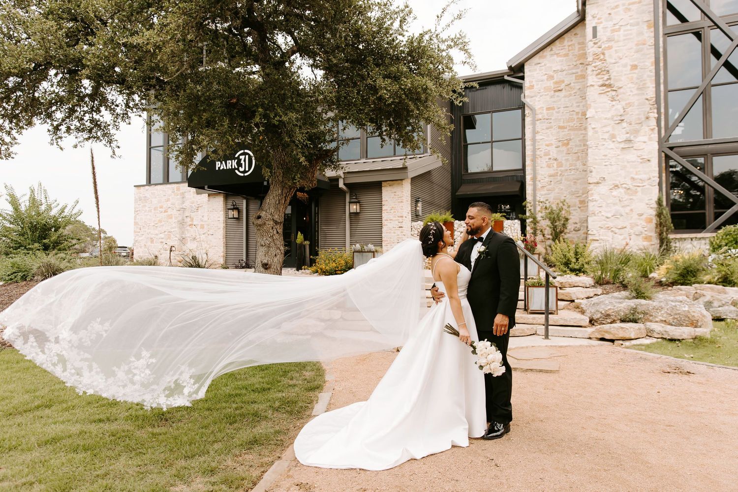 Bride and groom outside modern stone wedding venue with long flowing veil blowing in the wind, romantic outdoor wedding portrait.