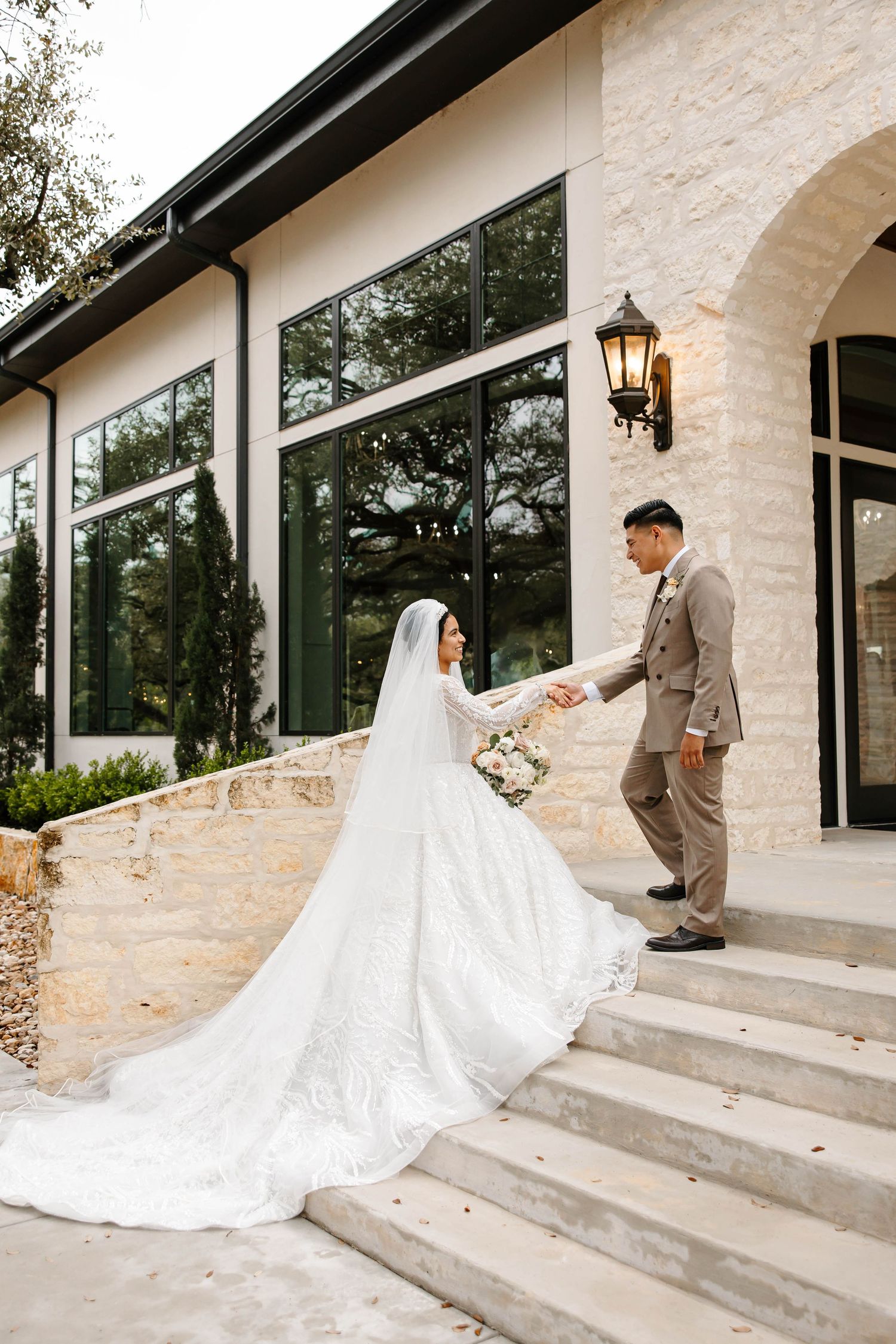 Bride and groom holding hands walking down stone steps outside a modern wedding venue with large windows and neutral stone exterior.