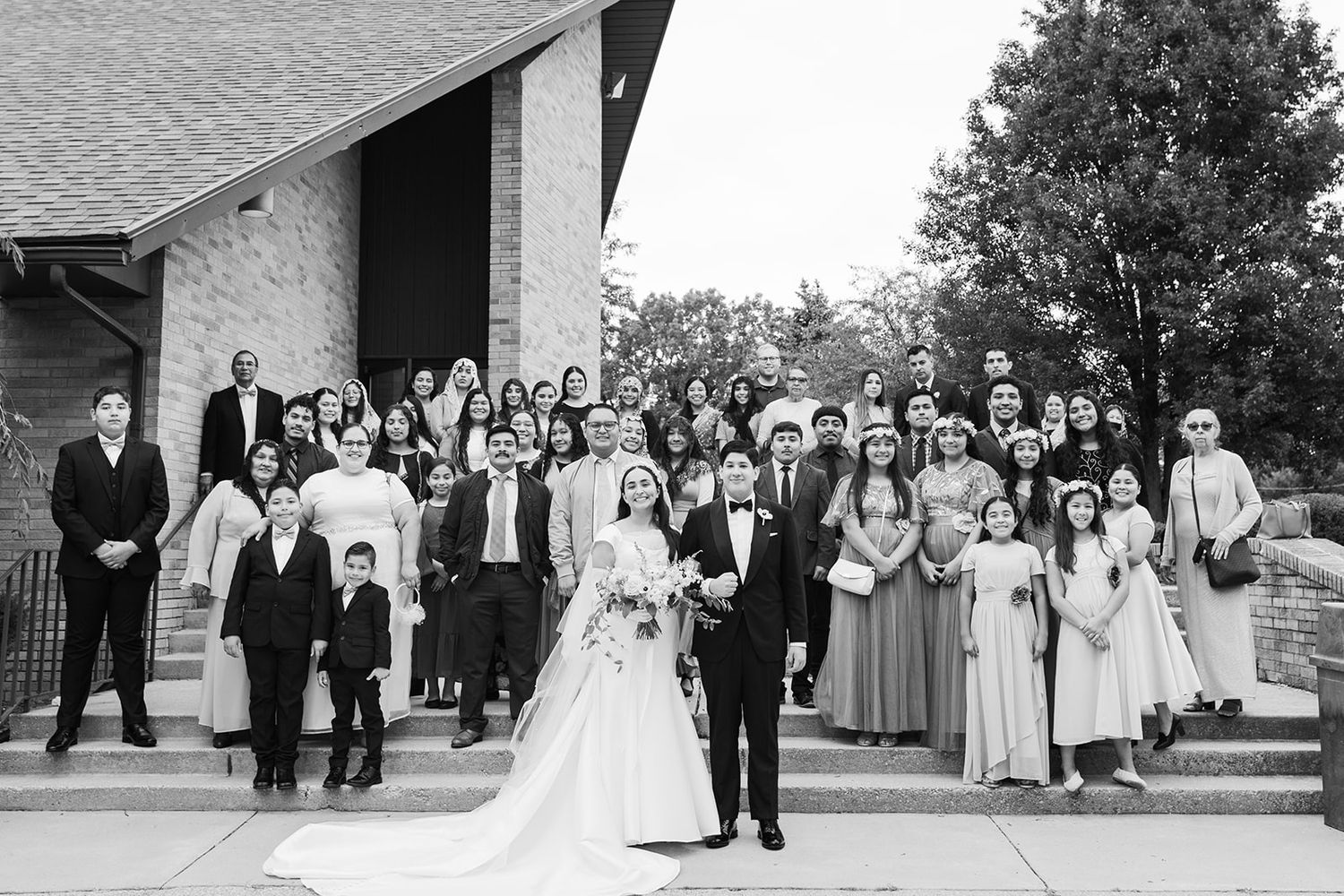 Large wedding party group portrait outside church with bride and groom centered and bridesmaids and groomsmen standing on steps.