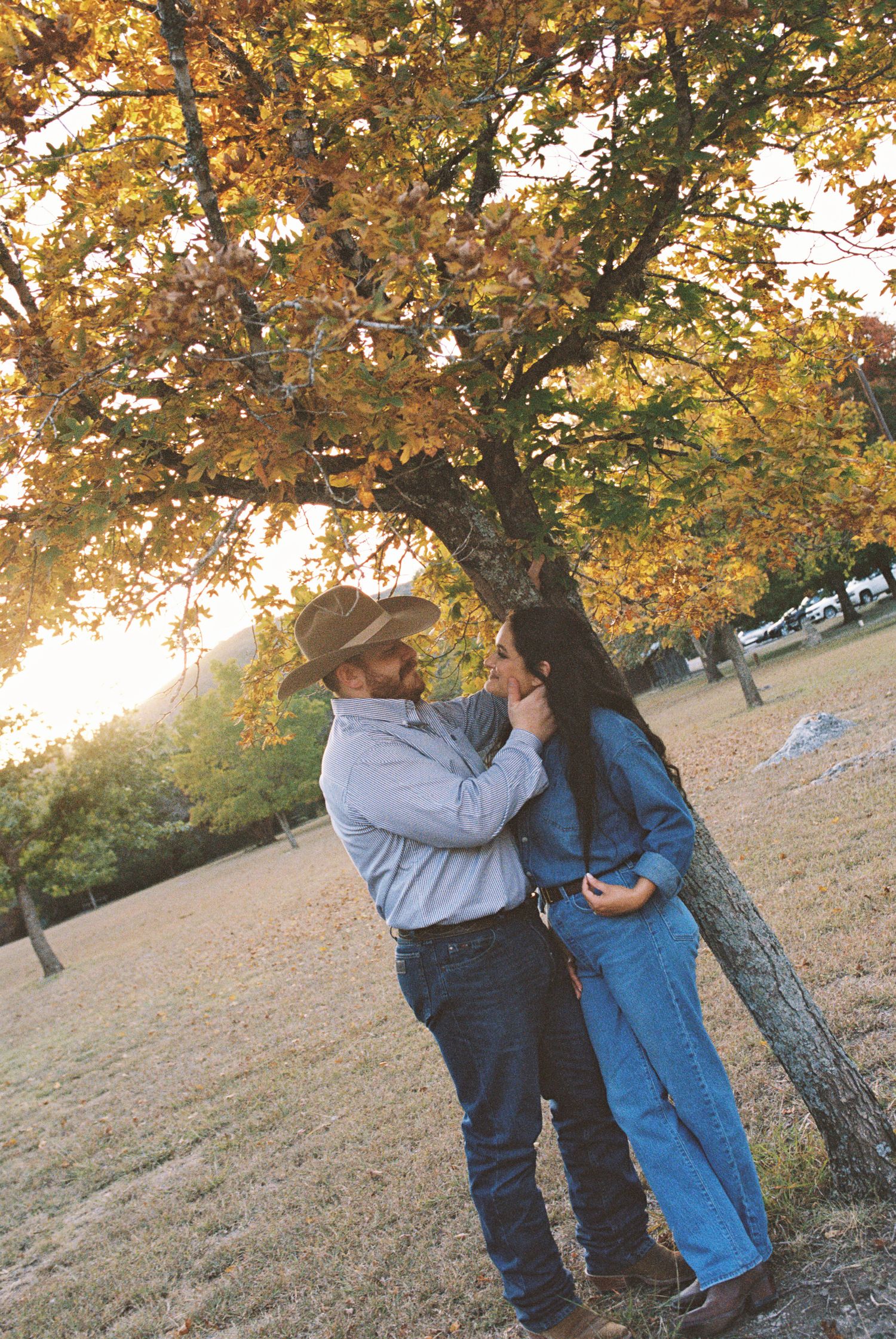 Couple under autumn trees, man in cowboy hat and woman in blue jeans outfit sharing a romantic outdoor moment.