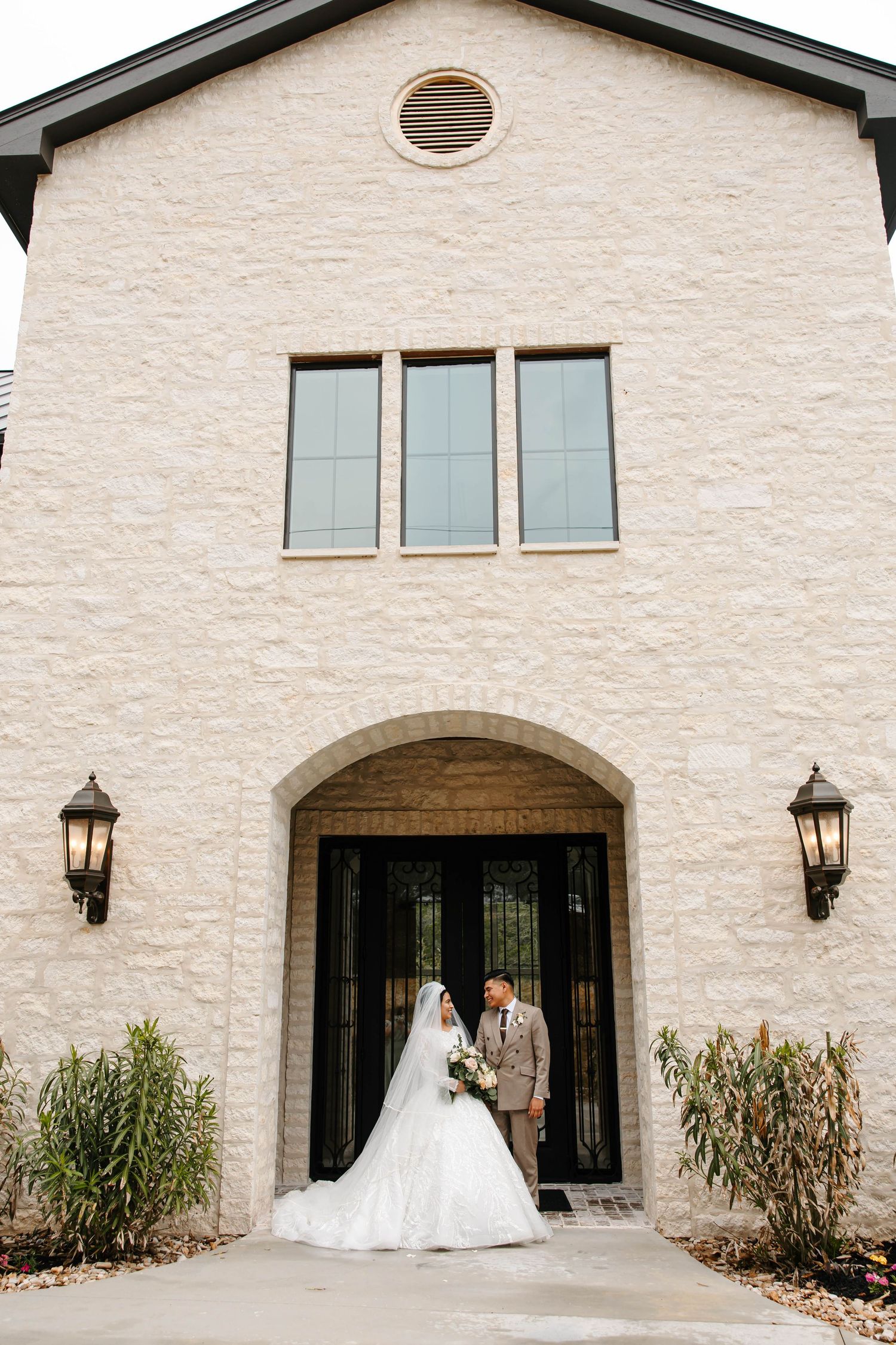 Elegant bride and groom standing in an arched doorway of a light stone chapel, classic wedding portrait with neutral architecture and soft