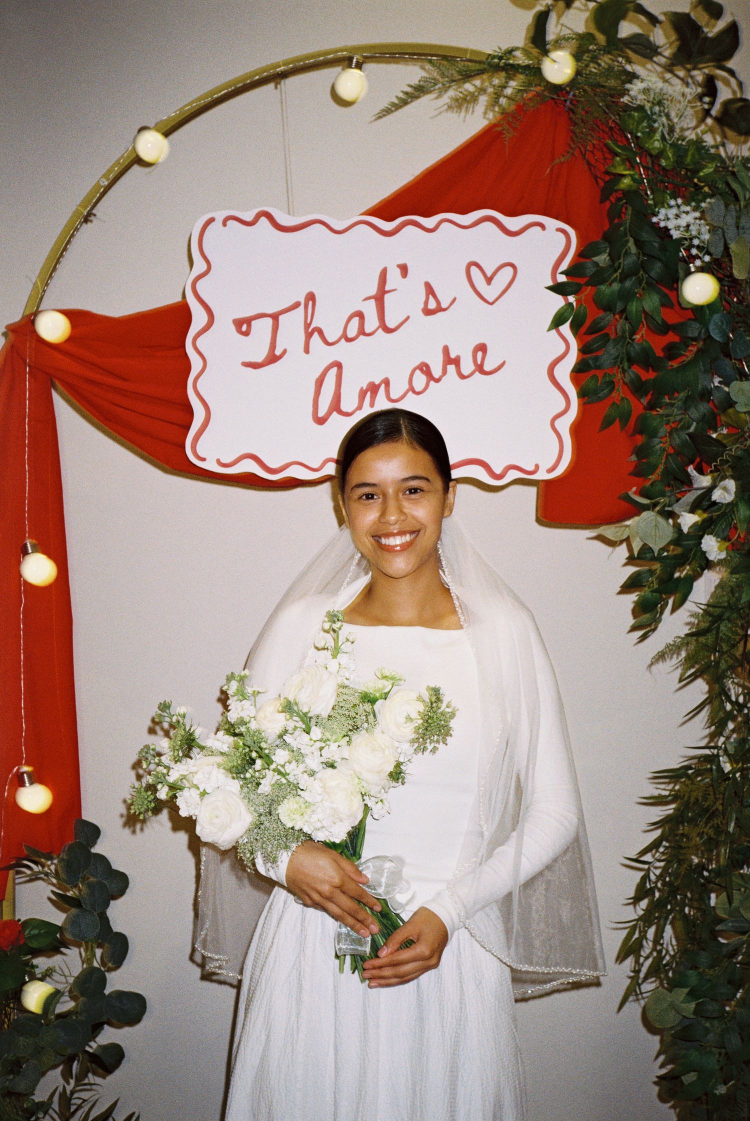 Bride holding bouquet under “That’s Amore” sign with red backdrop, elegant indoor wedding portrait.