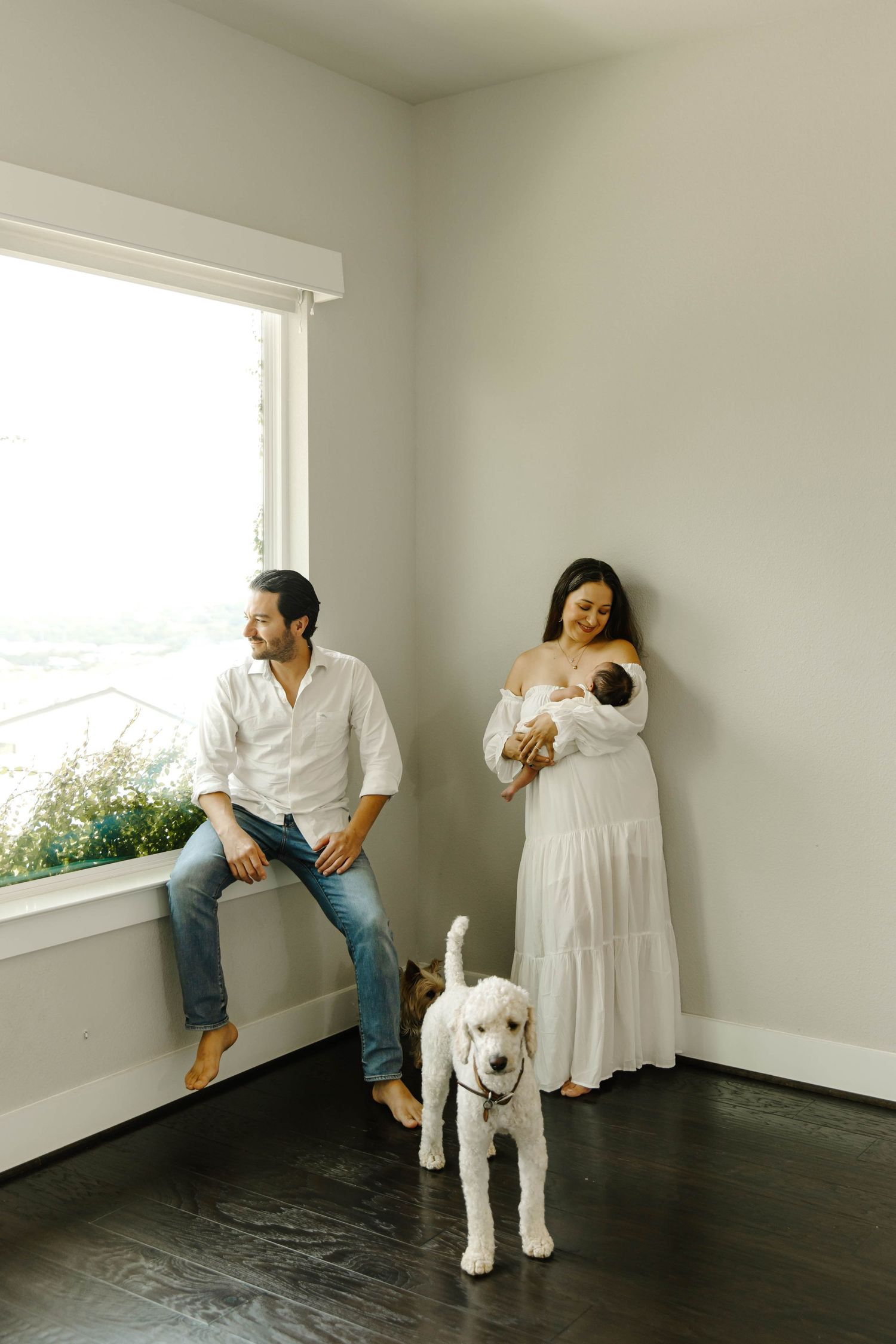 Bright lifestyle maternity photo of a couple in a minimalist room with large window, pregnant woman in white dress, partner sitting on windo