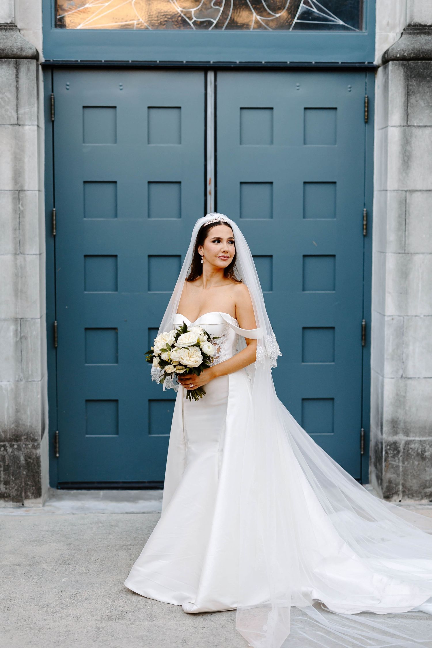 Bride in a classic white wedding gown holding bouquet standing in front of large blue double doors, modern bridal portrait.
