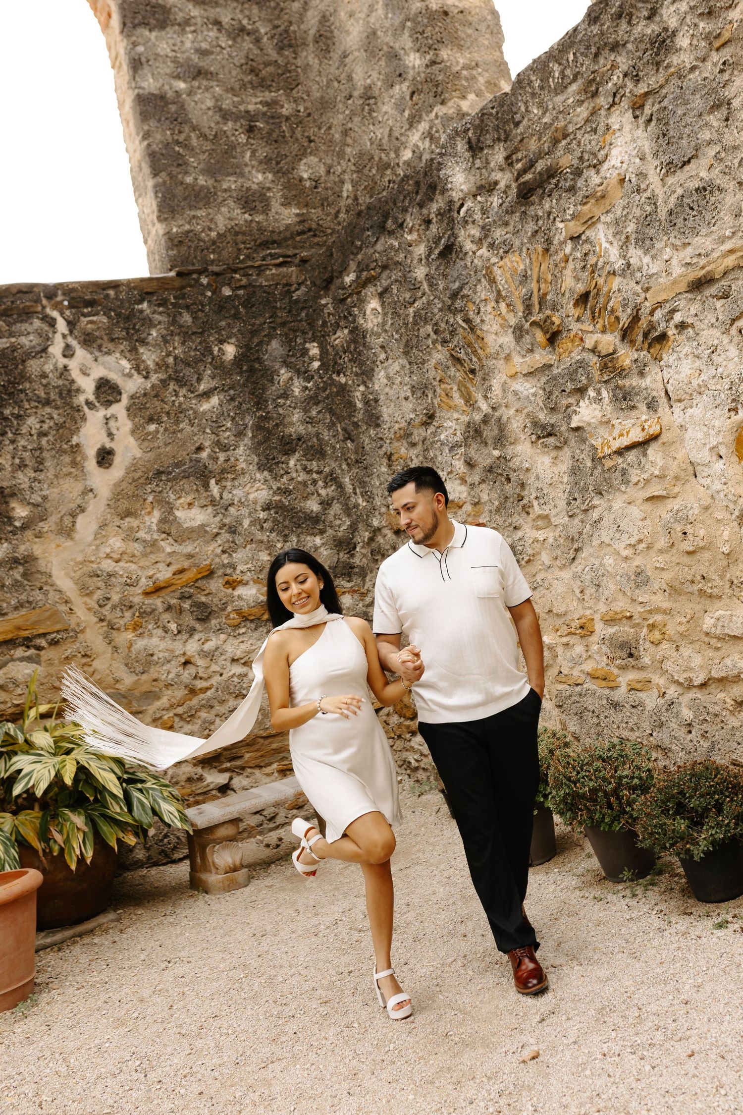 Elegant couple walking hand in hand beside historic stone wall, white dress and shirt, romantic travel photoshoot, soft neutral tones.