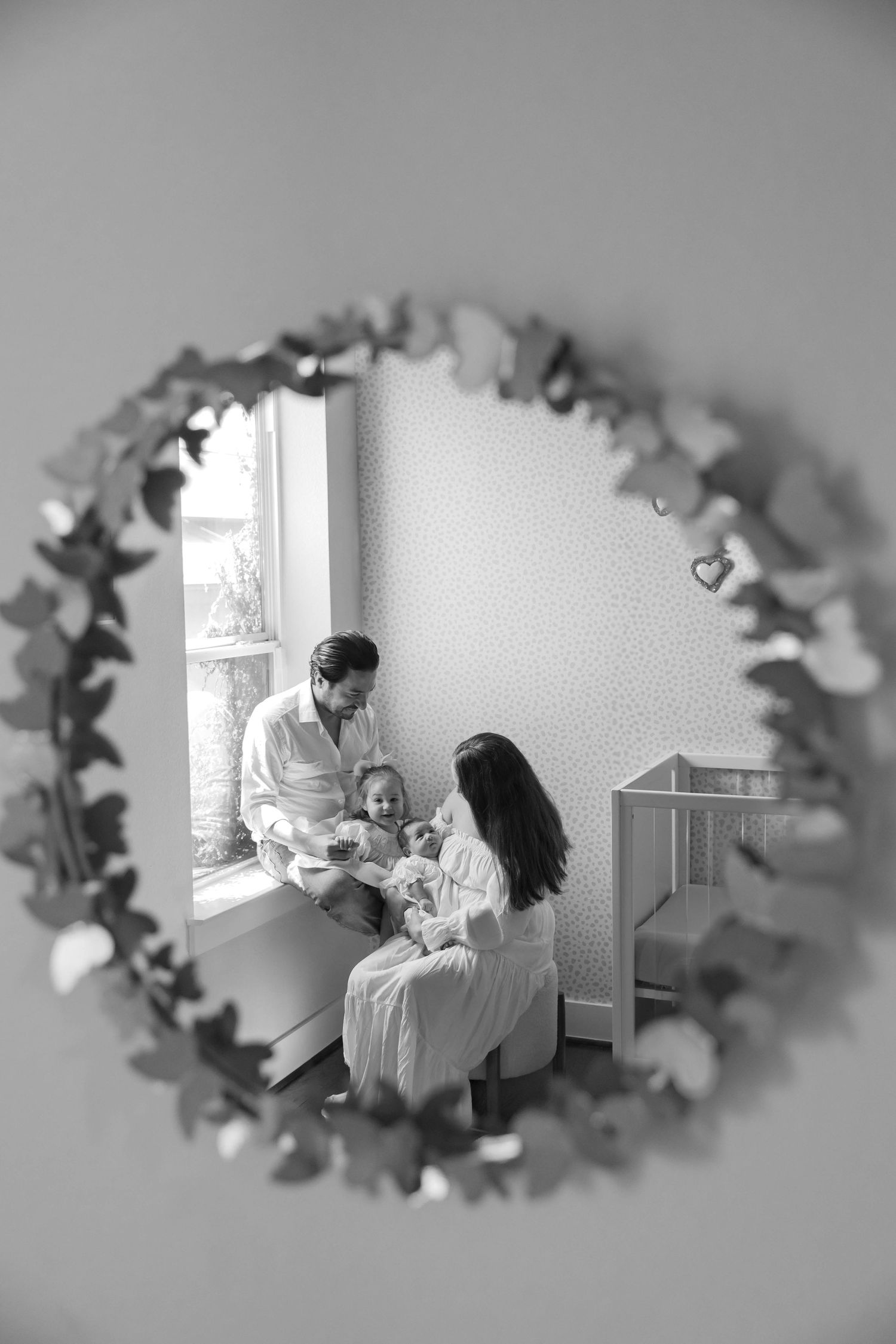 Black and white nursery scene framed through a round decorative mirror with leaf garland, parents sitting by a window holding their newborn