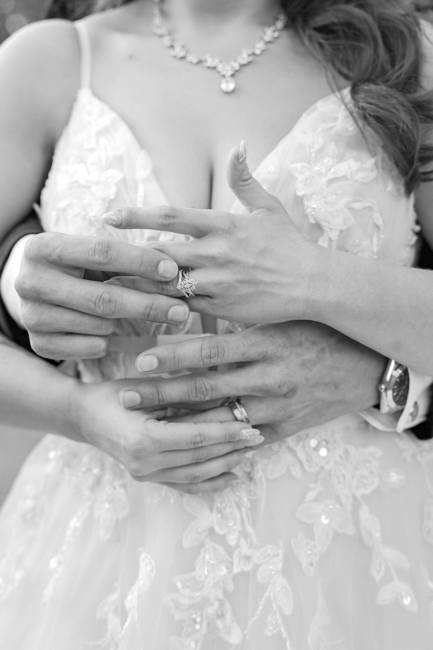 Black and white close-up of bride and groom hands showing wedding rings against lace wedding dress, intimate bridal detail photo.