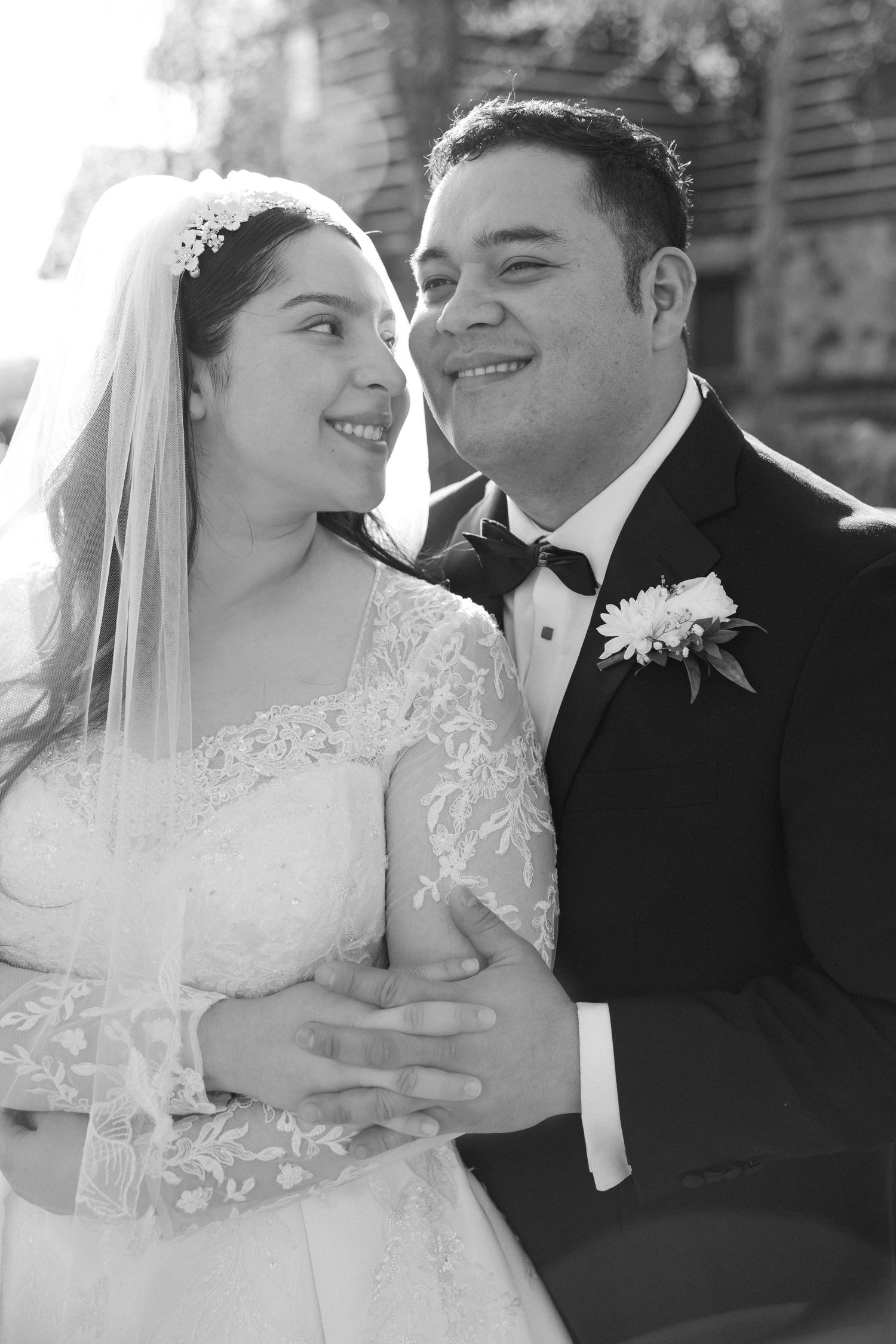 Black and white romantic portrait of newlyweds smiling at each other, bride in lace wedding dress and groom in tuxedo.