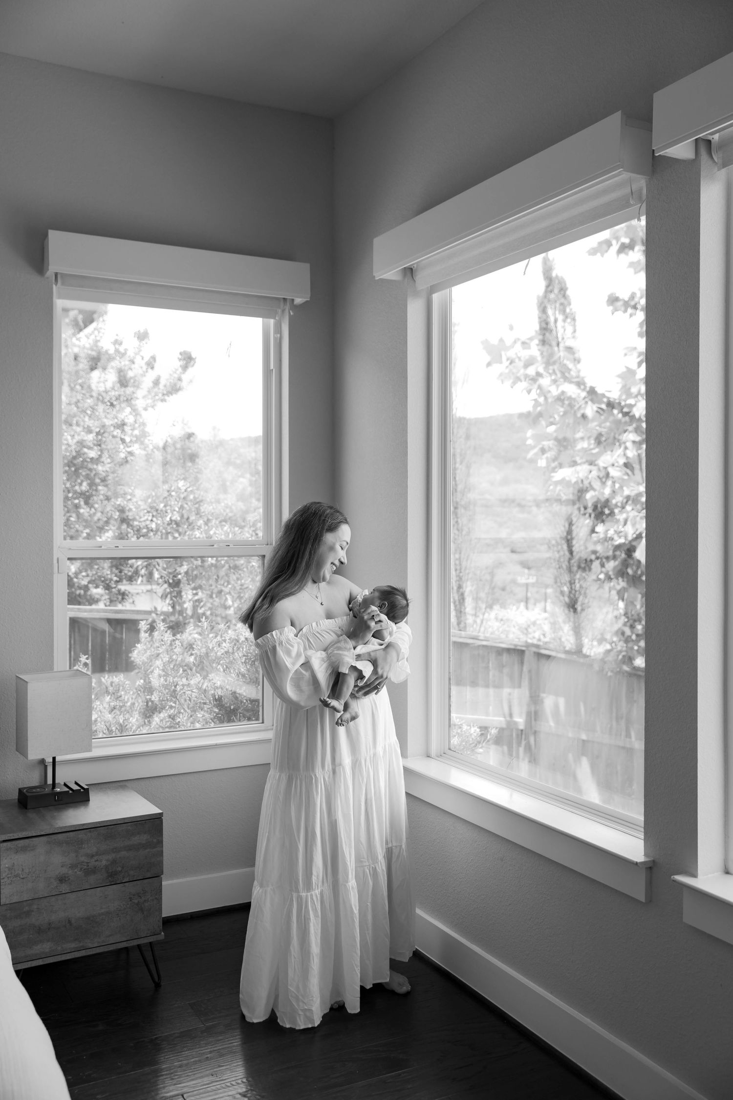 Black and white lifestyle portrait of a pregnant woman in a flowing white maternity dress standing by large bright windows, holding her baby