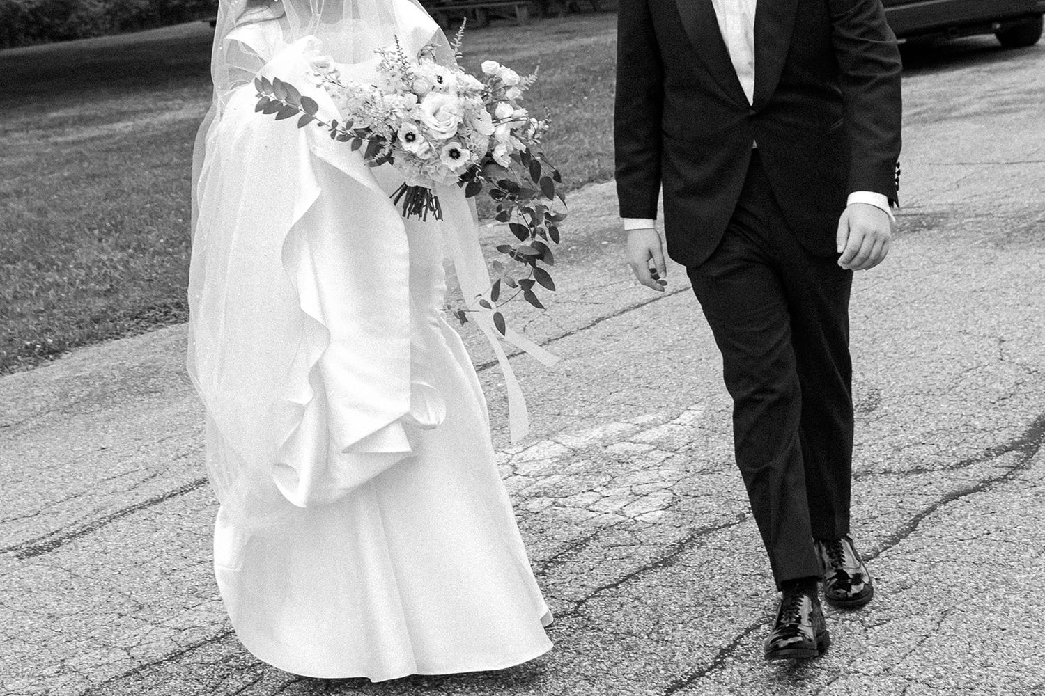 Black and white candid wedding photo of bride and groom walking together holding bouquet and hands along a city street.