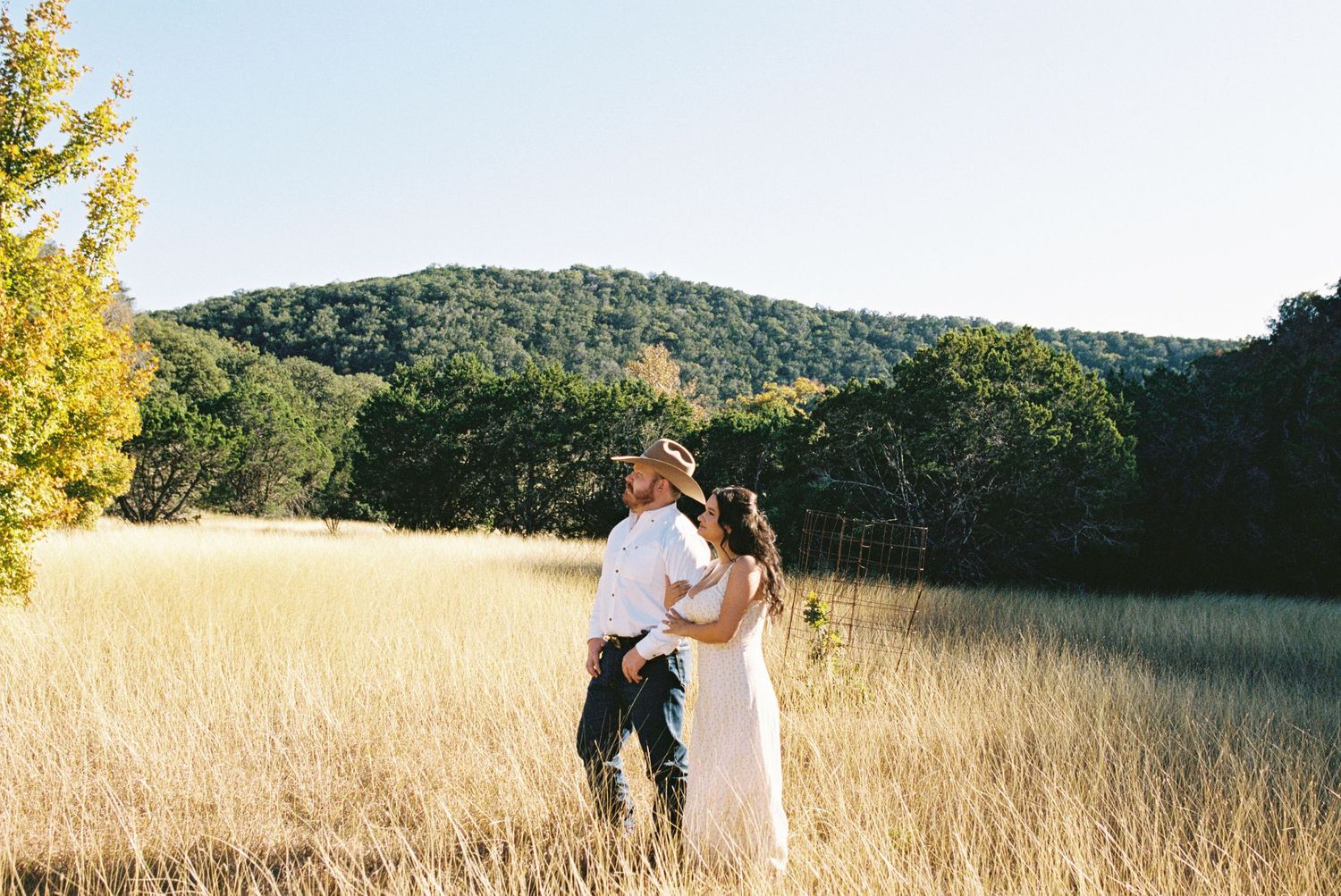 Couple standing among trees in fall foliage, embracing in soft sunlight, romantic woodland engagement portrait.